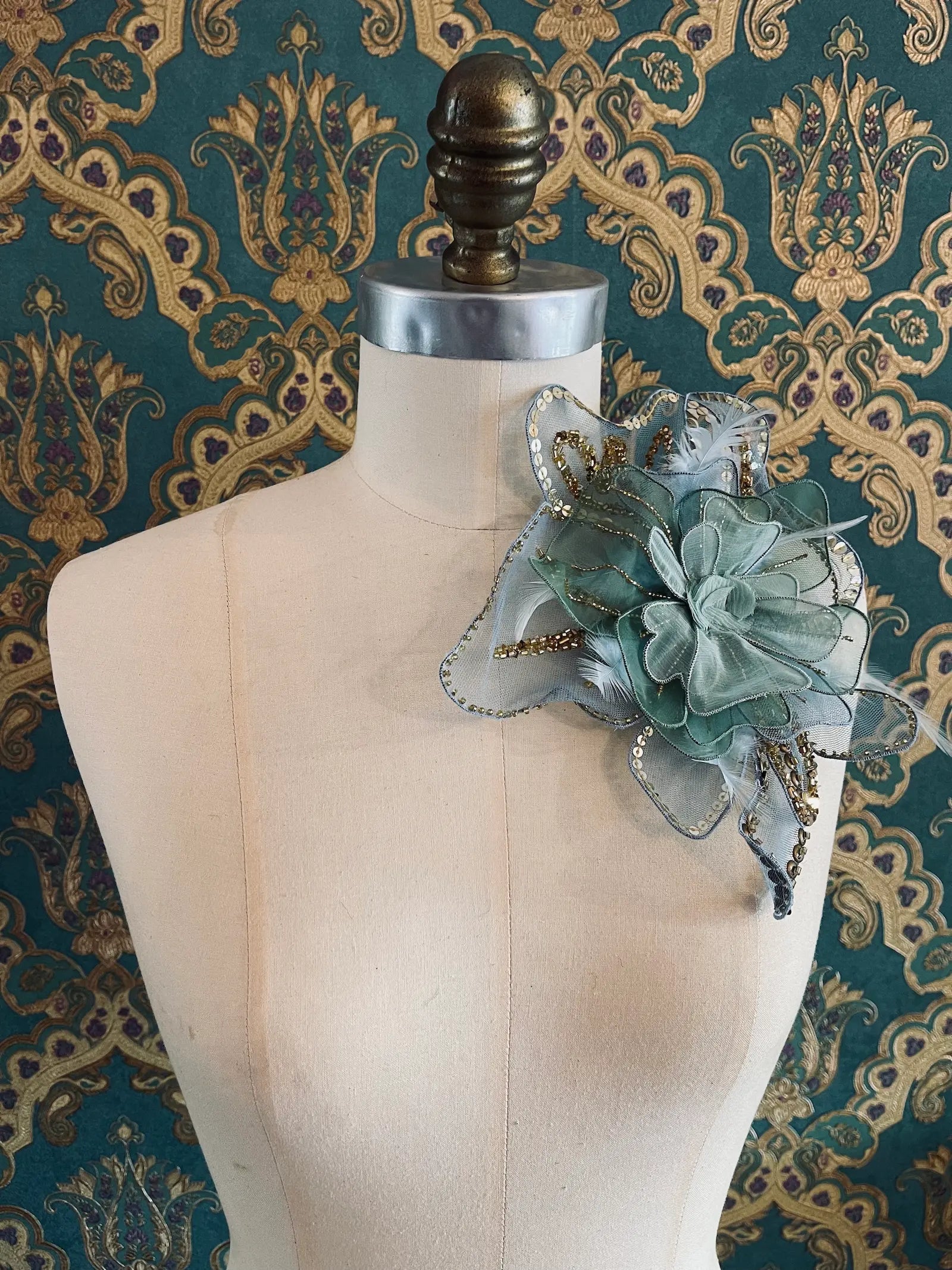 A Broochella Alstroemeraia large blue-green flower brooch  accessory with layered mesh flowers, sheer ribbon, and gold bead accents, displayed on a cream dress form against a teal and gold damask patterned background.