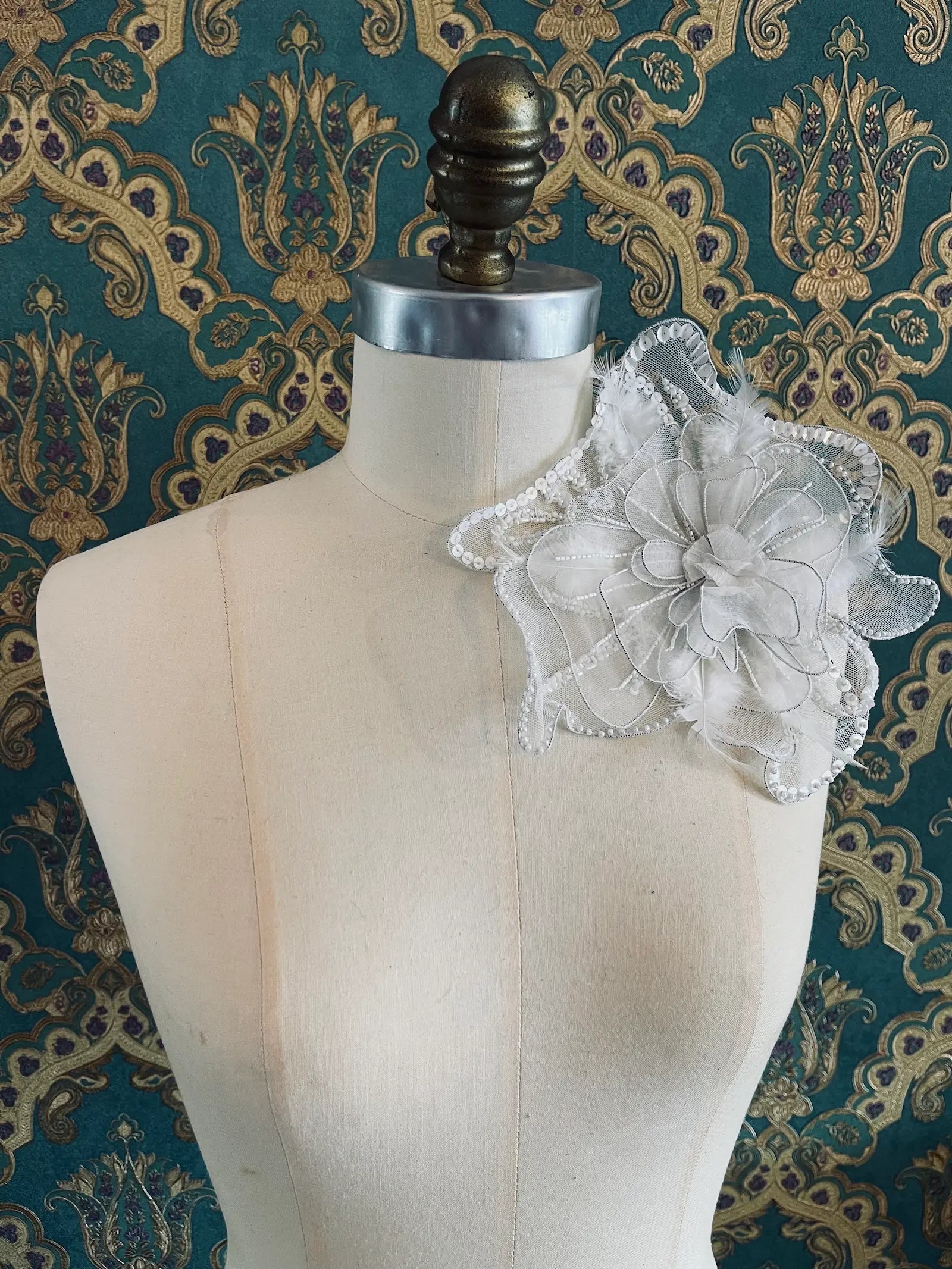 A Broochella Alstroemeraia large handmade white flower brooch  accessory with layered mesh flowers, sheer ribbon, and gold bead accents, displayed on a cream dress form against a teal and gold damask patterned background.