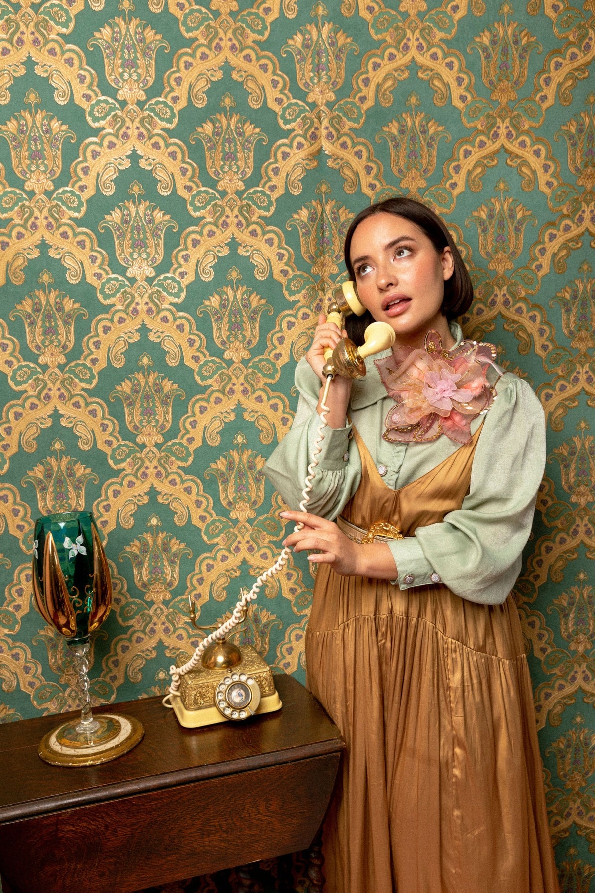 Broochella pink Alstroemeria flower brooch worn over a sage blouse and gold dress by a woman holding a vintage telephone against green and gold patterned wallpaper.