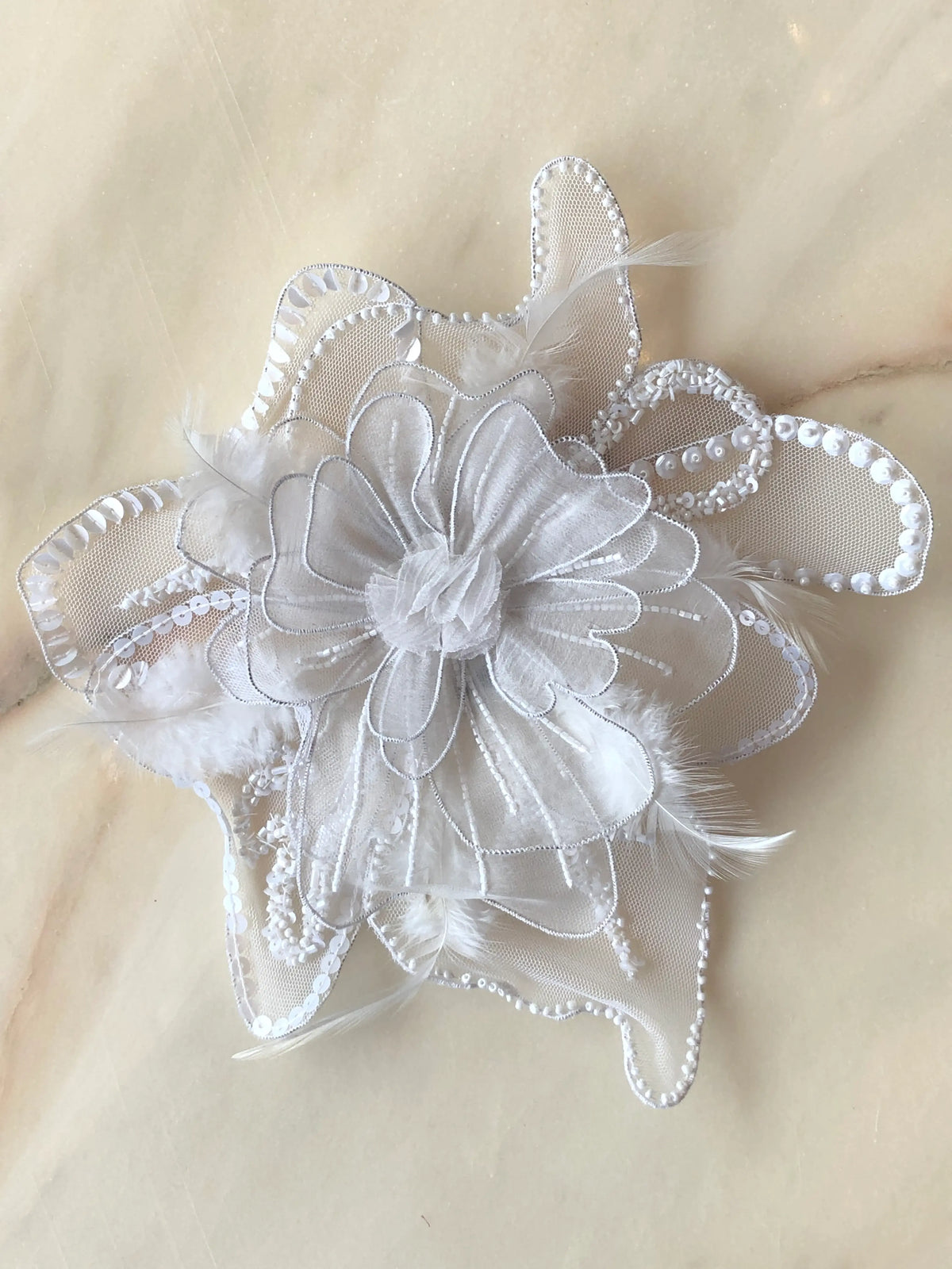 Broochella Alstroemeria Large white flower brooch, featuring layered white mesh petals embellished with white sequins, beads and delicate feather details, photographed on a pale cream marble background.