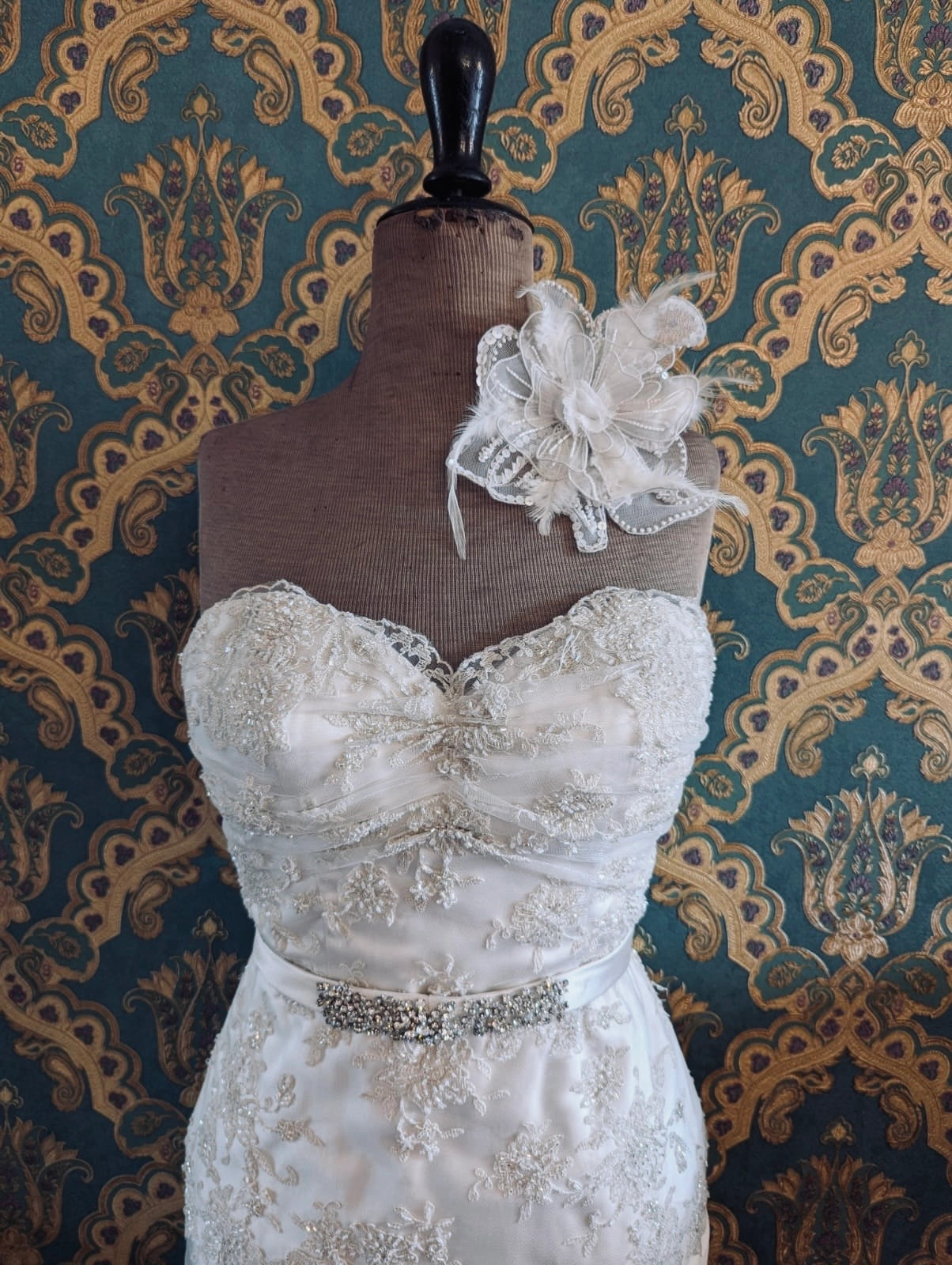 Broochella bridal alstromeria small white handmade beaded and feather flower brooch. Pinned on a mannequin that is wearing a white, beaded strapless wedding dress. The background is green and gold damask wallpaper. 