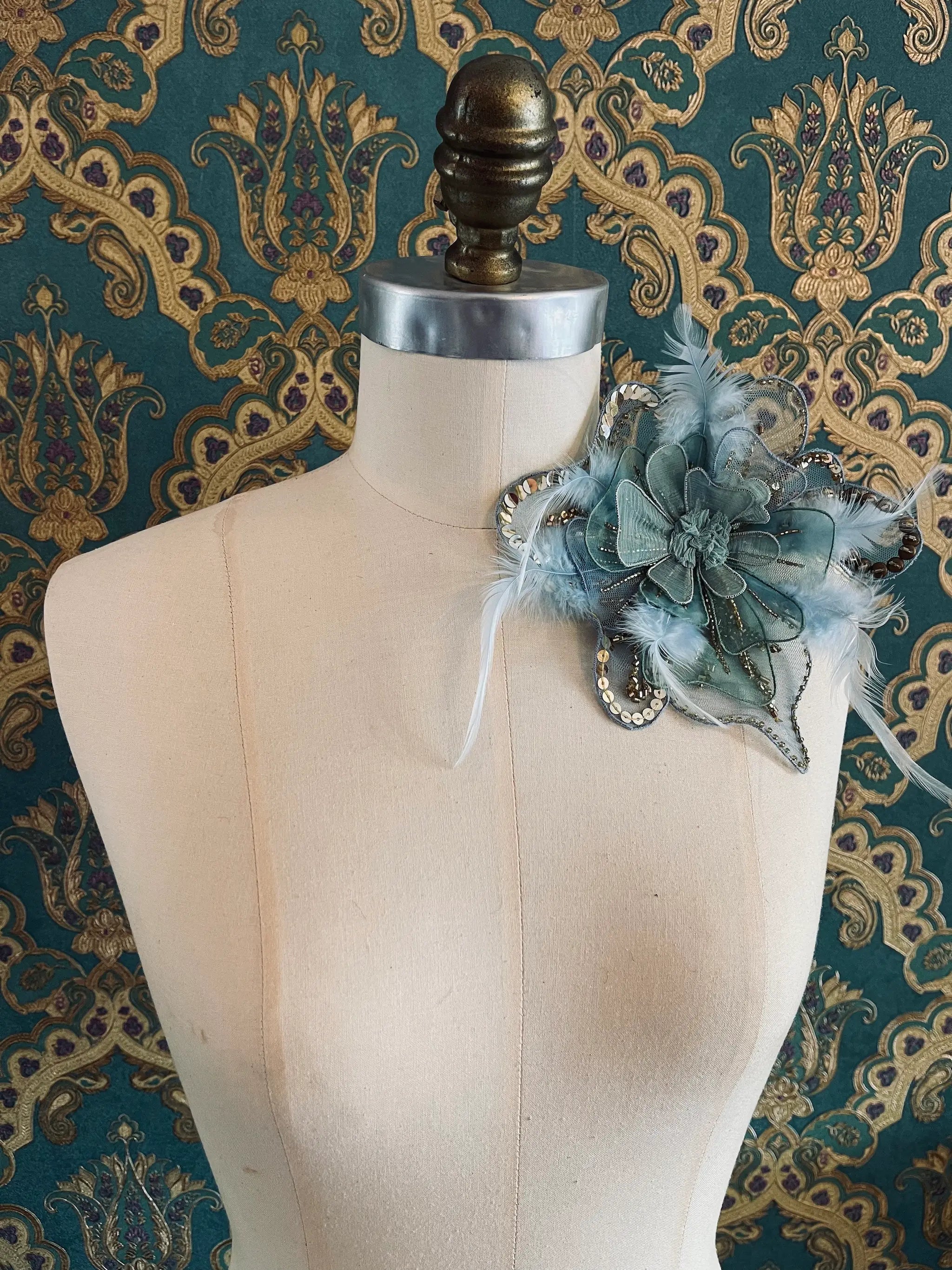 Broochella Alstromeria small hand beaded designer flower brooch in blue, green and gold. Pictured on a mannequin. The background is green and gold damask wallpaper