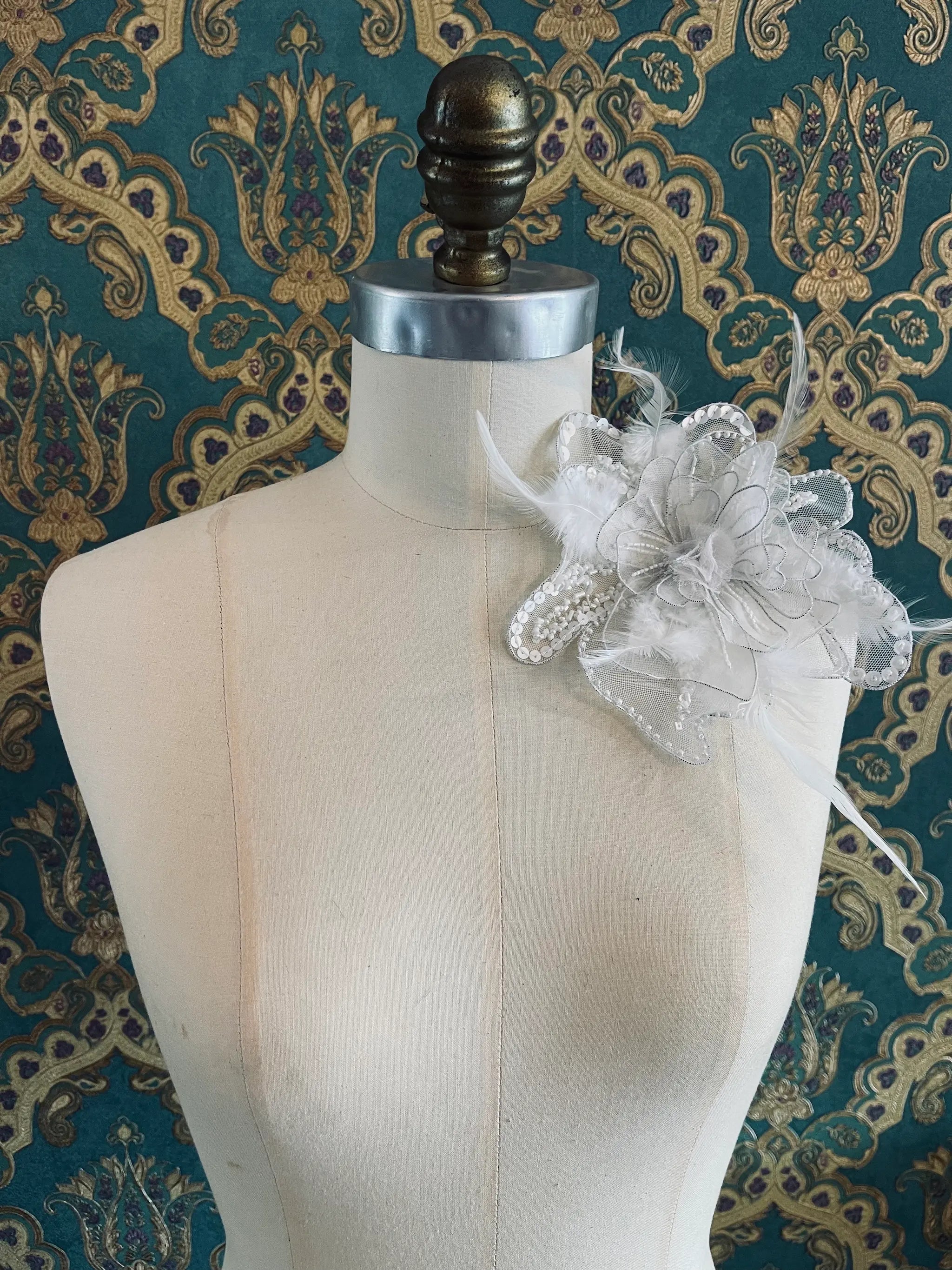 Broochella Alstromeria small hand beaded designer flower brooch in white. Pictured on a mannequin. The background is green and gold damask wallpaper