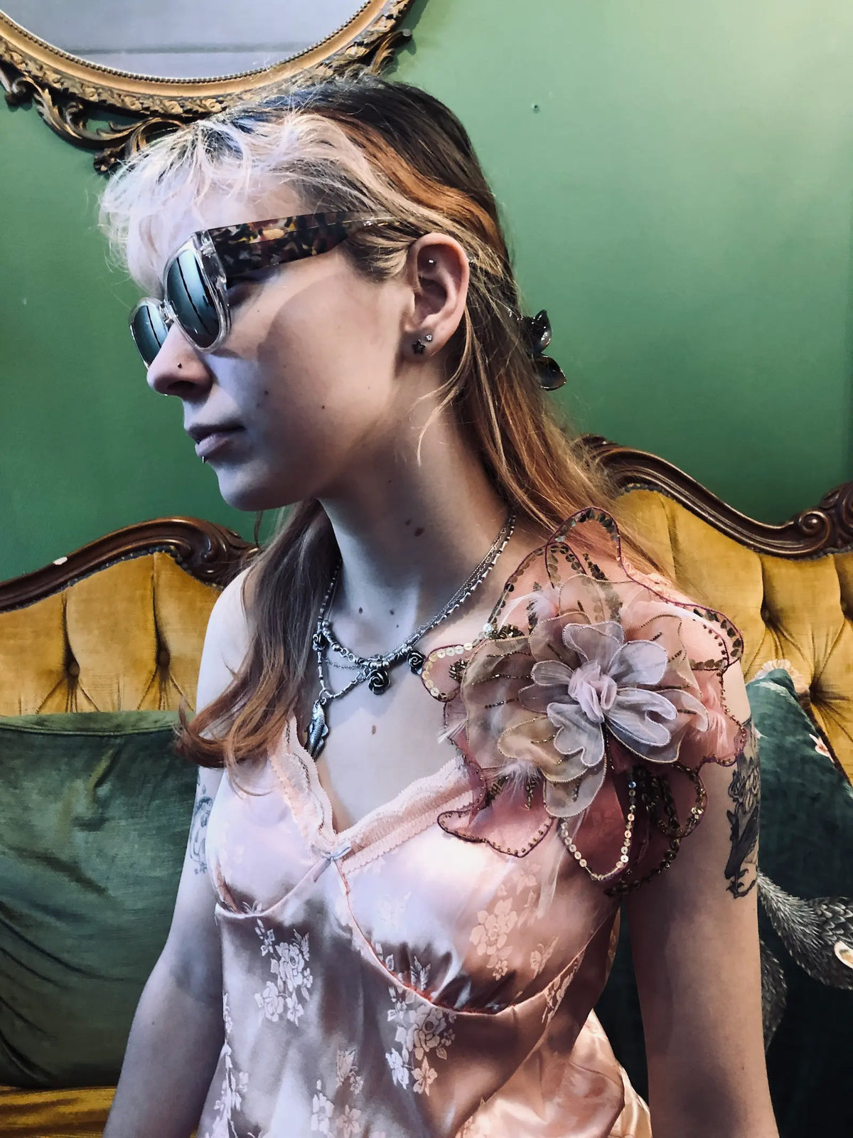 A person with glasses and tattoos poses against a dark green and gold damask patterned wallpaper, wearing a pale pink satin camisole and a Broochella Alstromeria brooch in pink