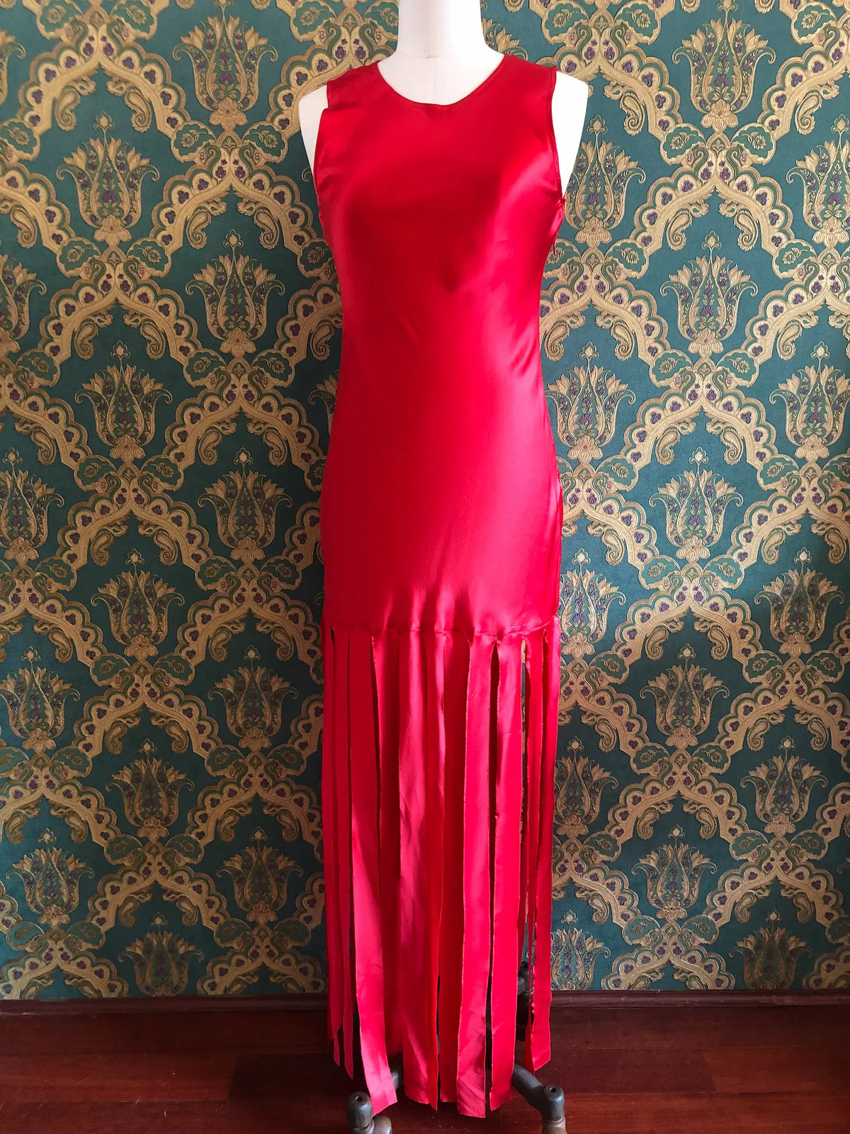 Broochella Amāre scarlet red silk maxi dress with a round neckline, sleeveless bodice and dramatic long panelled fringe, size AU 10. Pictured on a mannequin against green and gold damask wallpaper.