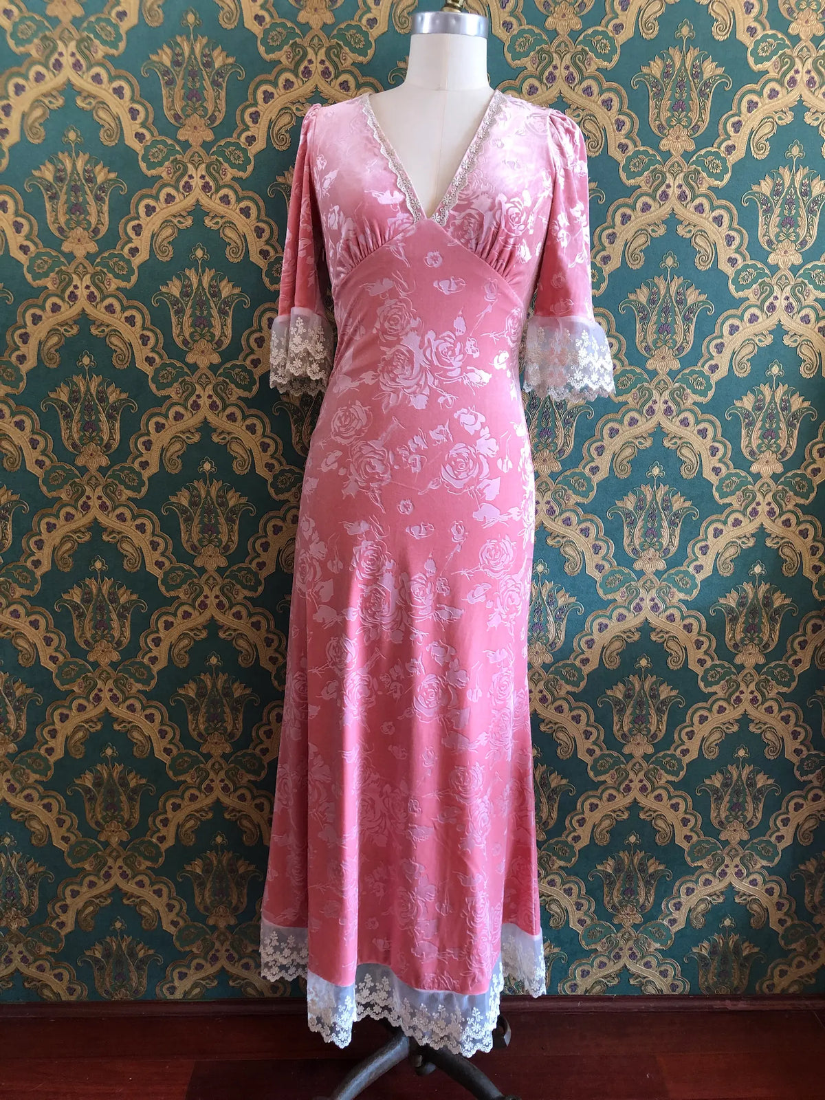 Broochella American Heiress Gwyneth maxi dress in Dusty Rose Pink with embossed floral velvet, a deep V-neckline and ivory lace trim, size Medium. Pictured on a mannequin against green and gold damask wallpaper.