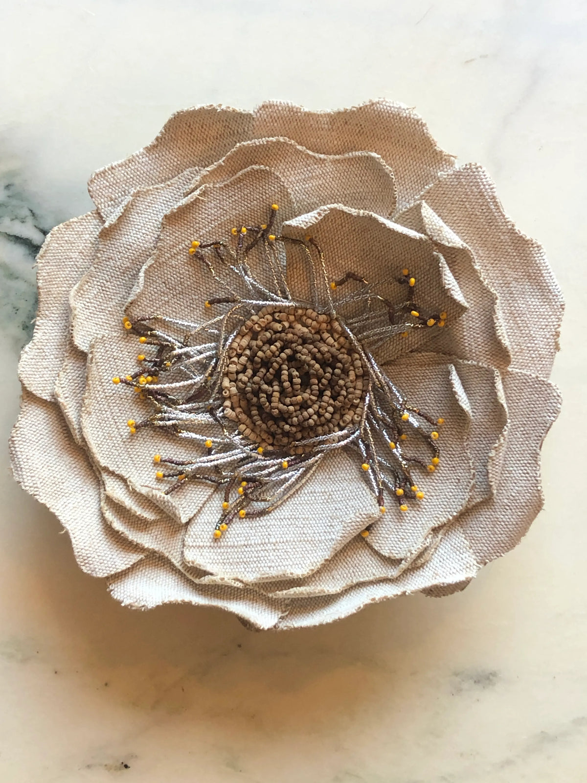 Broochella Anemone Bridal Flower Brooch featuring layered ivory linen petals, a hand-beaded centre and delicate metallic embroidered details with tiny yellow bead accents, photographed on a cream marble background.