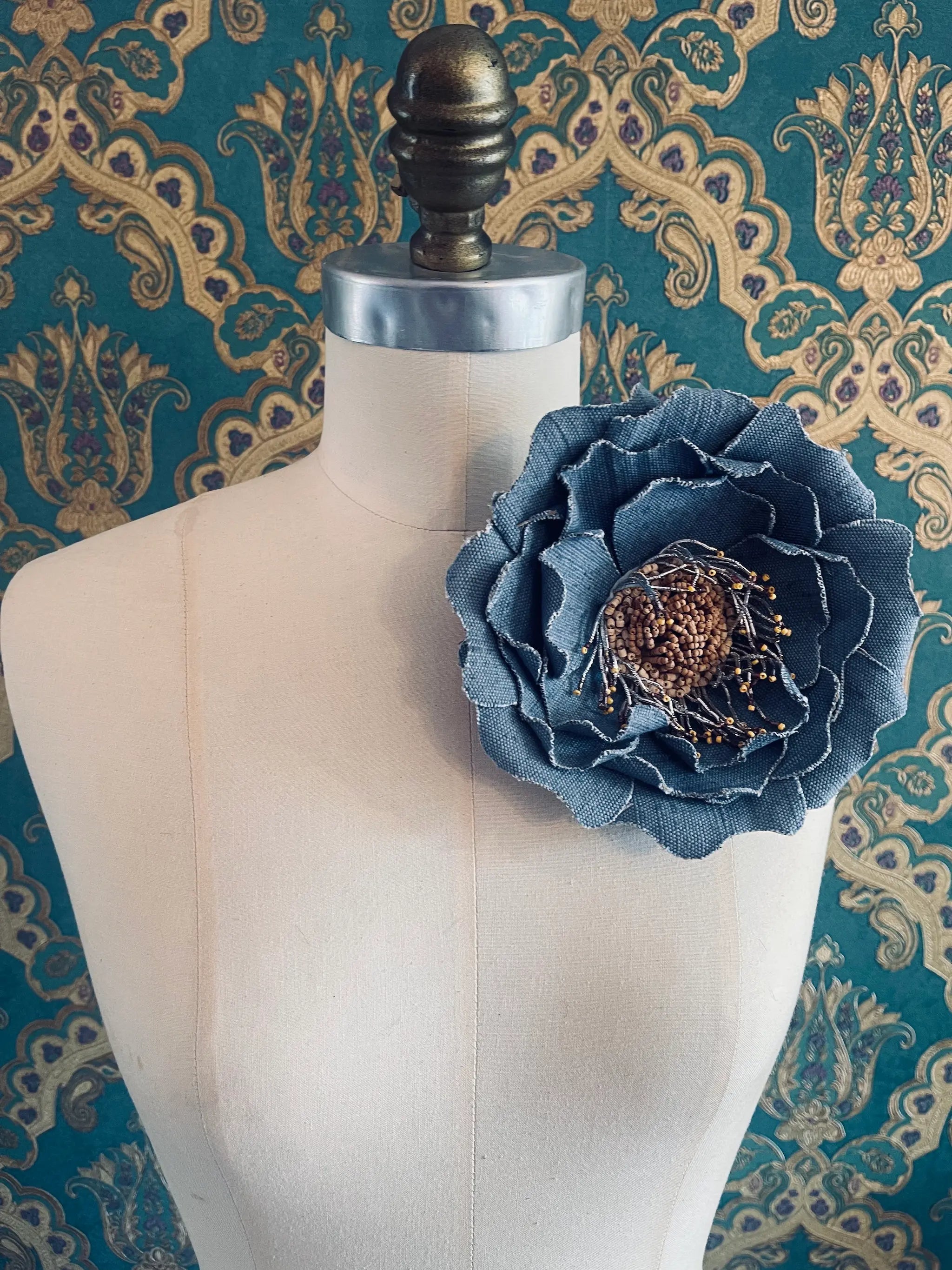 Broochella Anemone large handmade jute fabric flower brooch in blue. Styled on a mannequin. The background is green and gold damask wallpaper