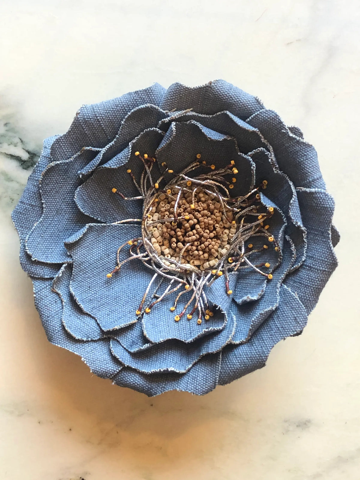 Broochella Anemone Flower Brooch Pin in soft denim blue, featuring sculptural layered fabric petals with a hand-beaded centre and delicate metallic stamen detailing on a cream marble background.