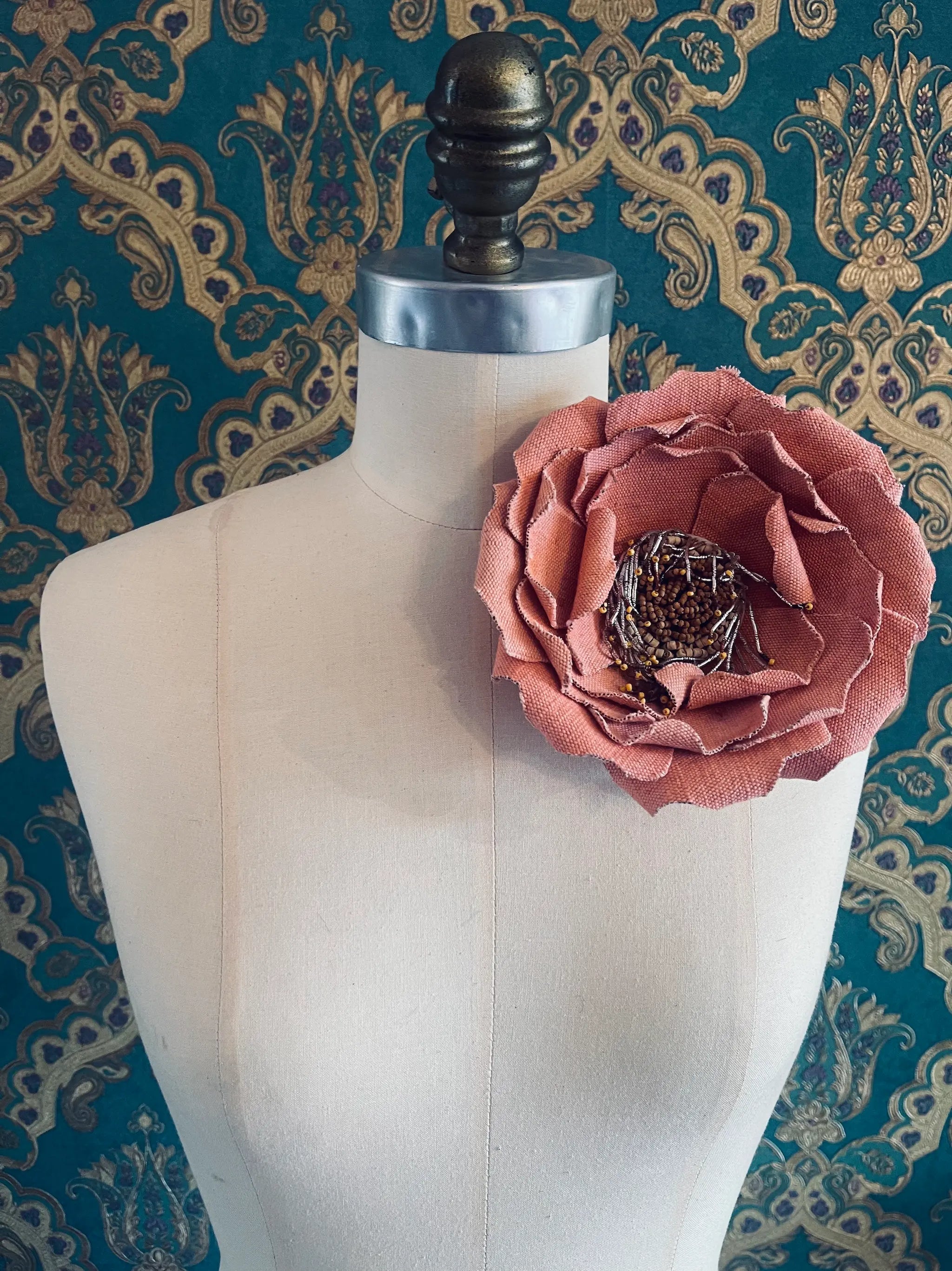 Broochella Anemone large handmade jute fabric flower brooch in pink. Styled on a mannequin. The background is green and gold damask wallpaper