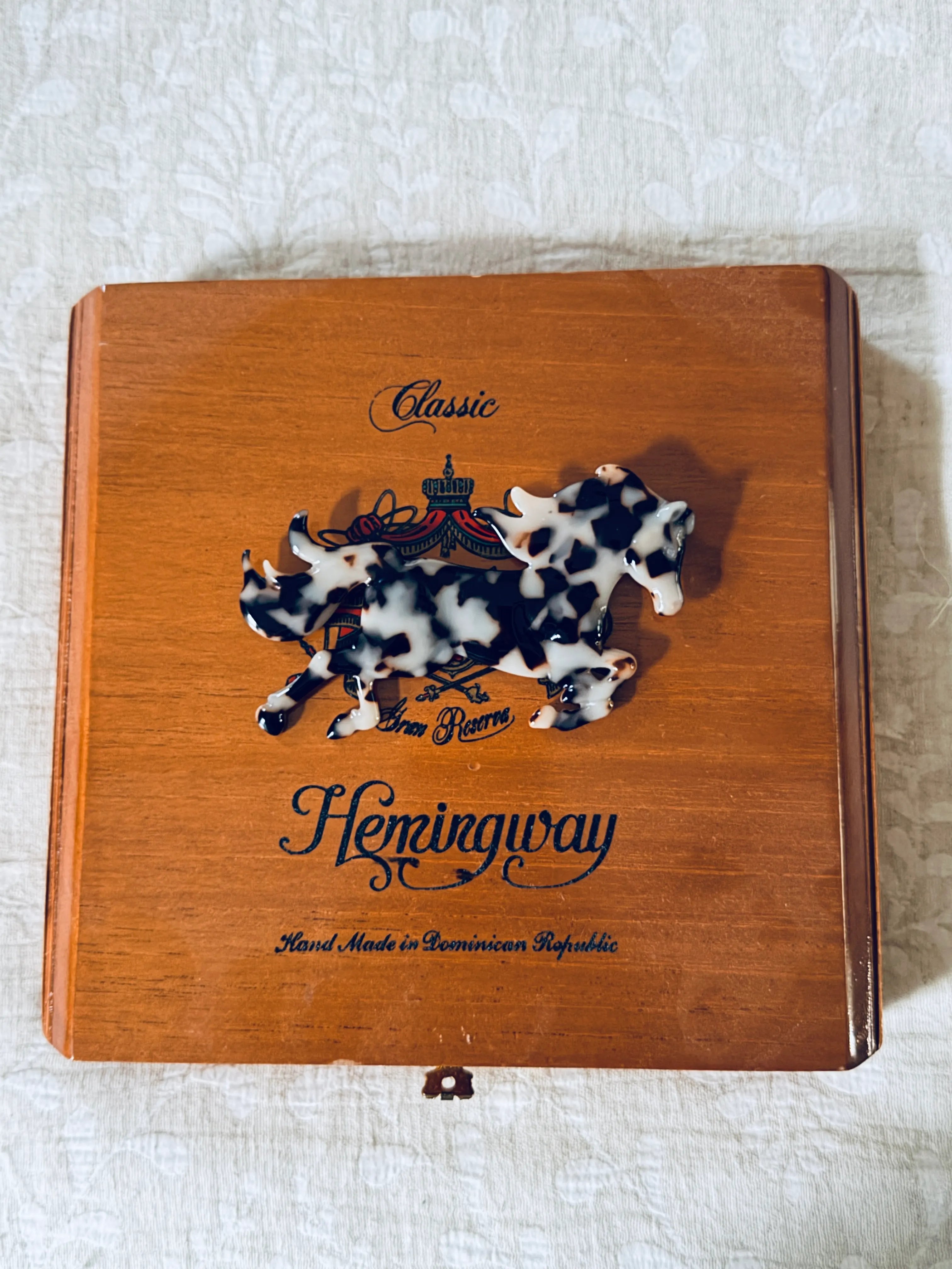 Broochella animalia horse acetate figural brooch. Pictured on a wooden cigar box. The background is beige damask fabric