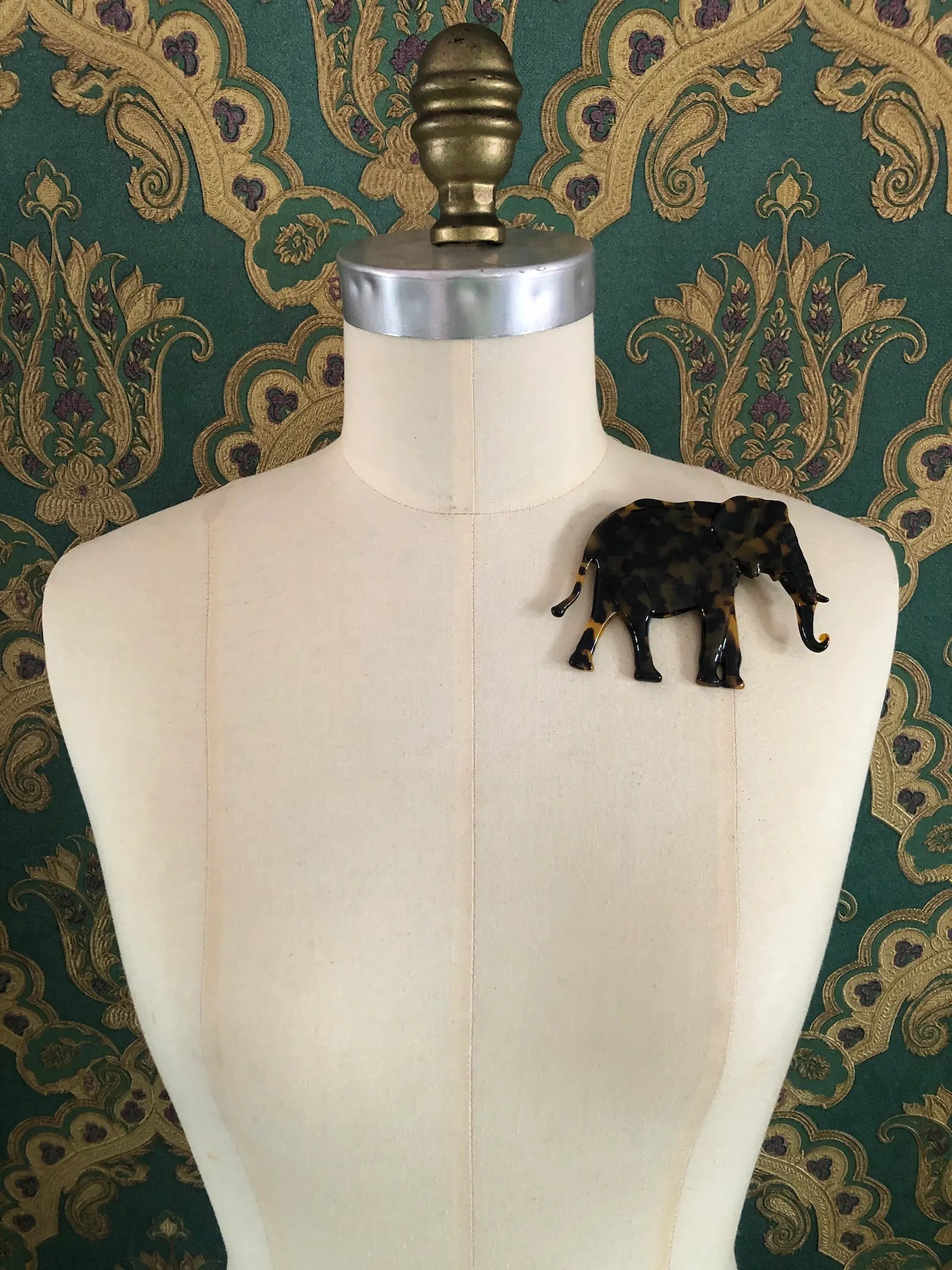 Broochella Animalia brown, figural  hand crafted, cellulose acetate elephant brooch in brown tortoiseshell. It is pinned to a mannequin The background is green and gold damask wallpaper.