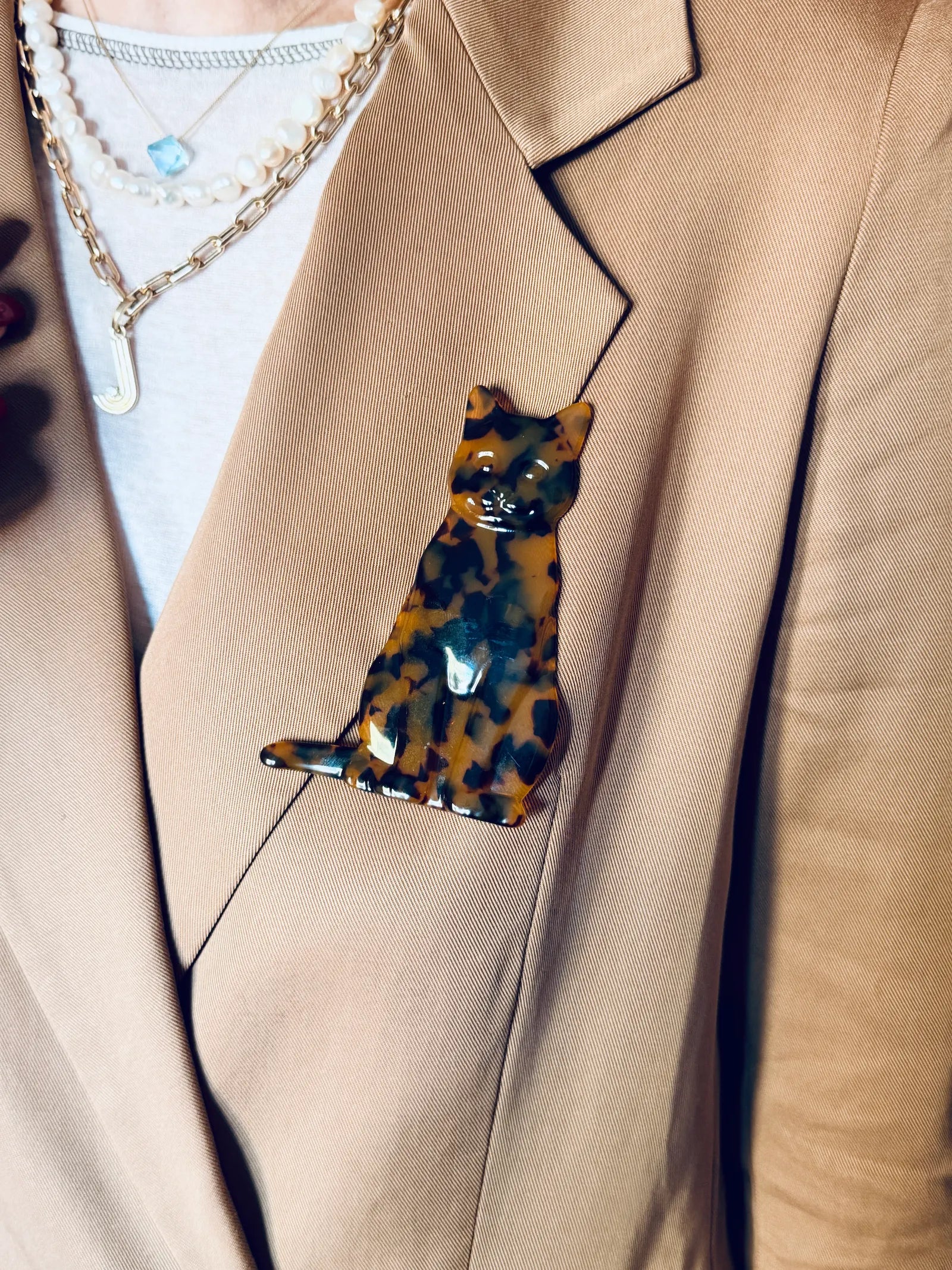 A Broochella Animalia brown tortoiseshell cat-shaped brooch pinned to a beige blazer over a white shirt with layered gold necklaces.
