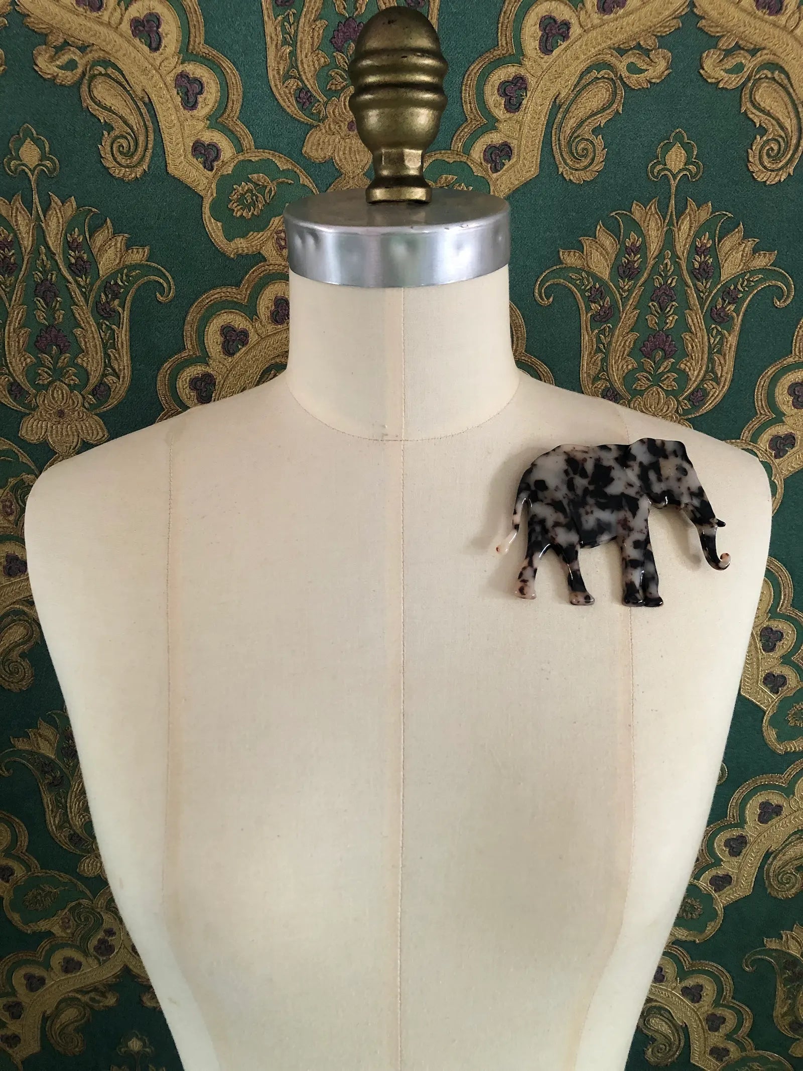 Broochella Animalia brown, figural  hand crafted, cellulose acetate elephant brooch in beige tortoiseshell. It is pinned to a mannequin The background is green and gold damask wallpaper.