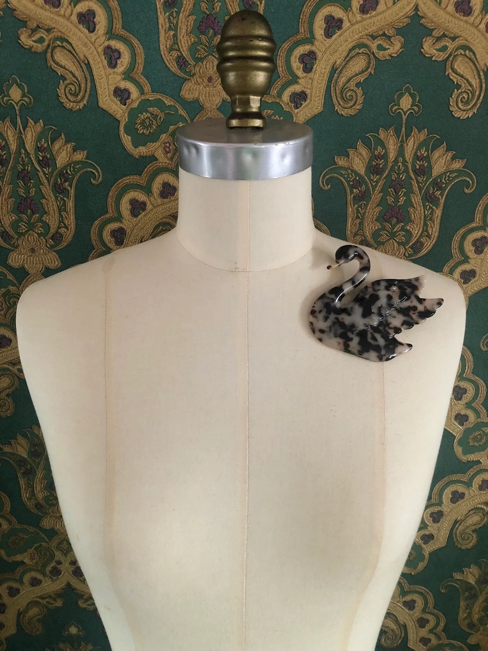 Broochella Animalia brown, figural  hand crafted, cellulose acetate swan brooch in beige tortoiseshell. It is pinned to a mannequin The background is green and gold damask wallpaper.