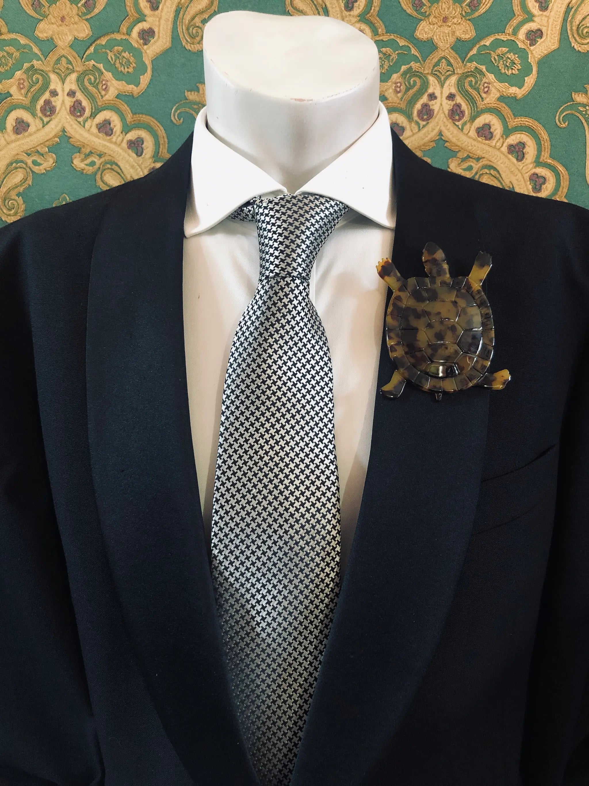 Broochella Men's brown Animalia tortoiseshell tortoise shaped brooch. It is made from Premium Italian cellulose tortoiseshell acetate. It is presented on a men's mannequin that is dressed in a black suit. The background is green and gold damask wallpaper. 