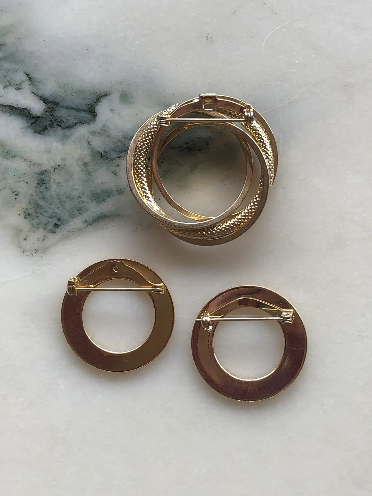 Broochella Anneaux d’Or Vintage Brooch Trio back view showing the gold-tone construction and traditional pin fastenings, photographed on a white marble background with soft grey-green veining.