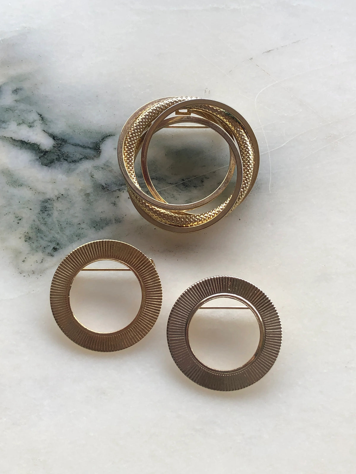 Broochella Anneaux d’Or Vintage Brooch Trio featuring one intertwined gold-tone ring brooch and two textured circular brooches, photographed on a white marble background with soft grey-green veining.