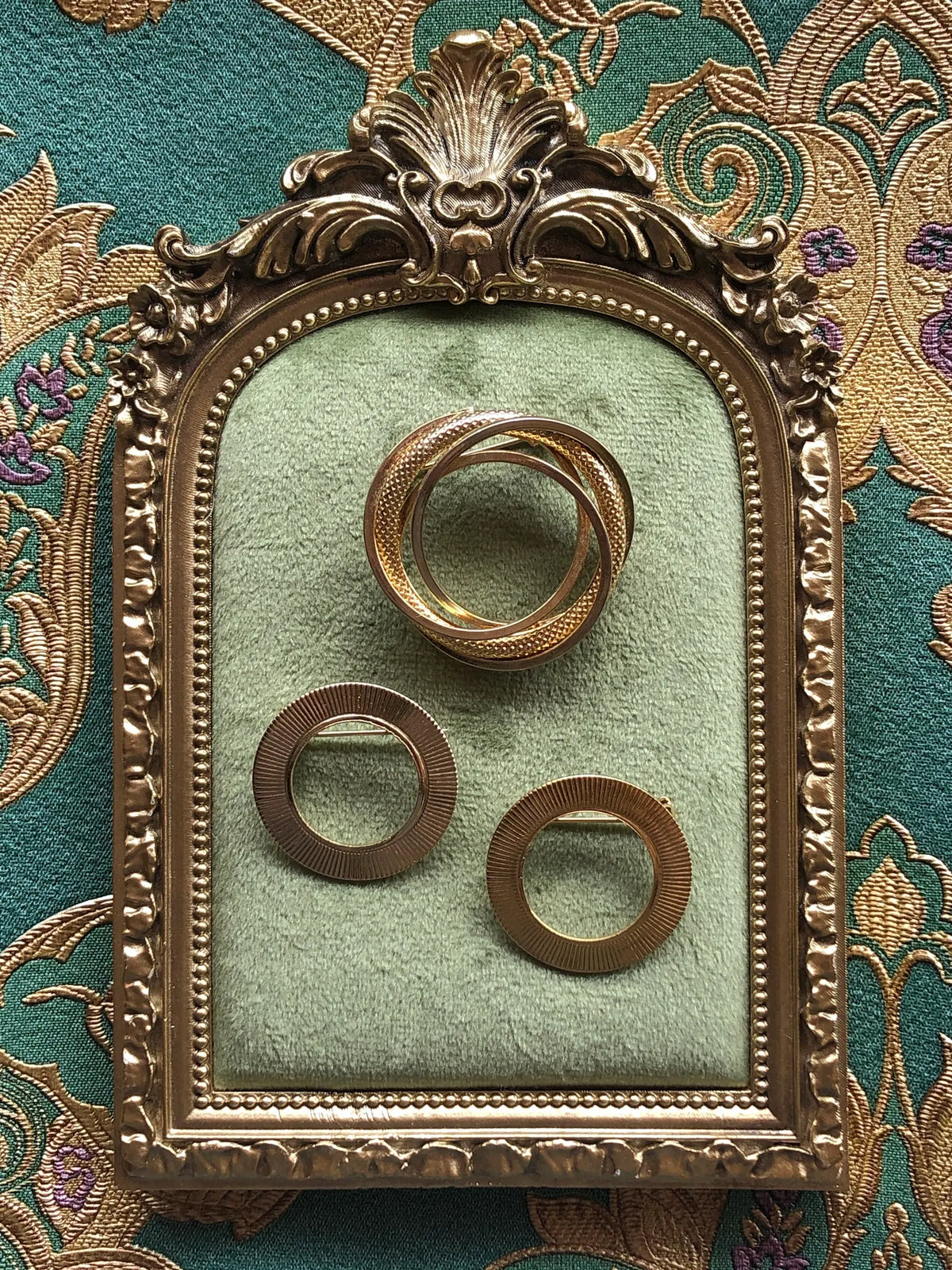 Broochella Anneaux d’Or Vintage Brooch Trio featuring three sculptural gold-tone circular brooches with textured and intertwined ring designs, displayed on sage green velvet in an ornate antique gold frame against green and gold patterned fabric.