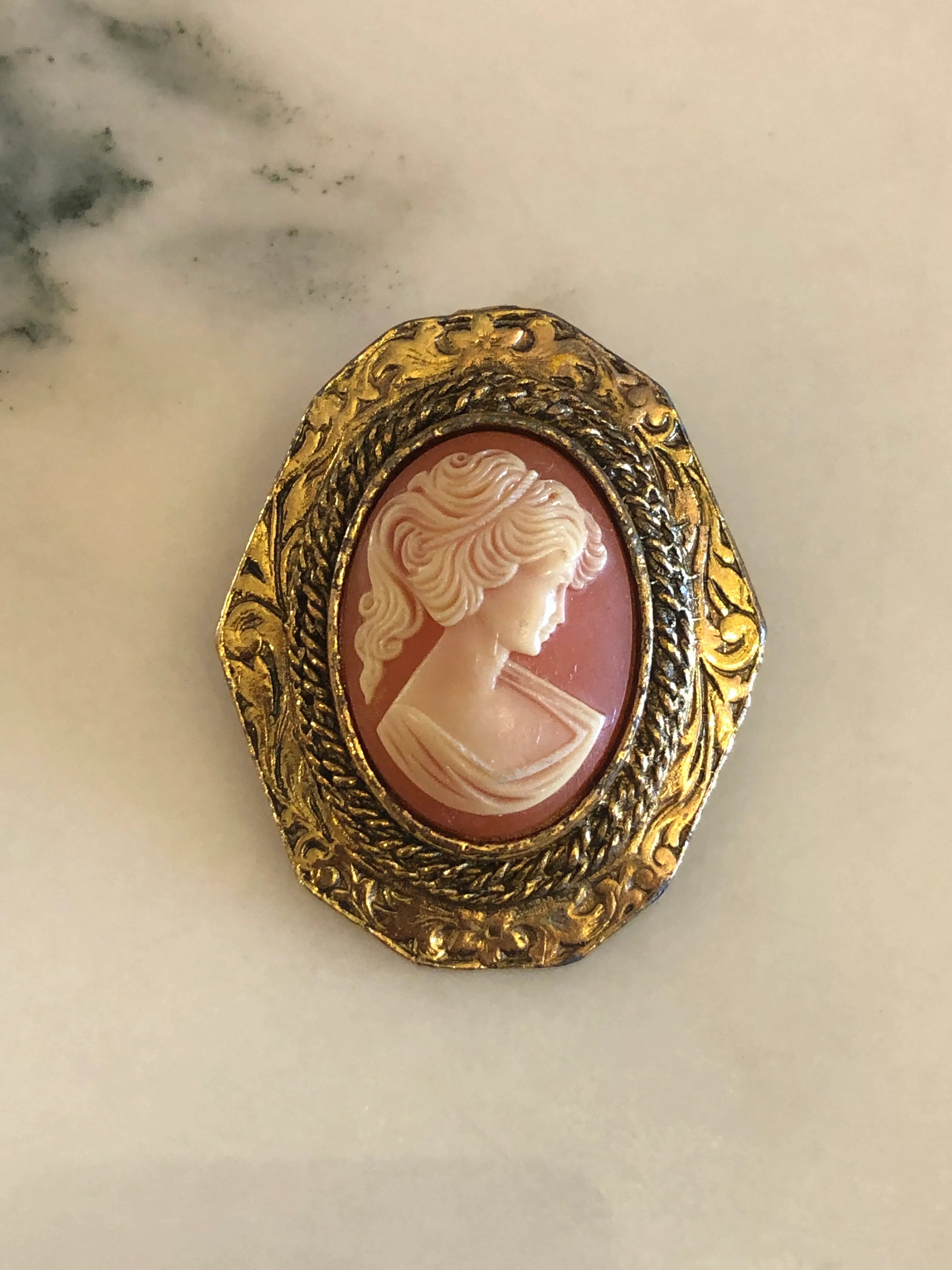 An Broochella vintage Antico peri  oval brooch featuring a carved pink and cream shell cameo of a woman's profile, set in an ornate textured gold-tone frame, displayed on a light marble surface.