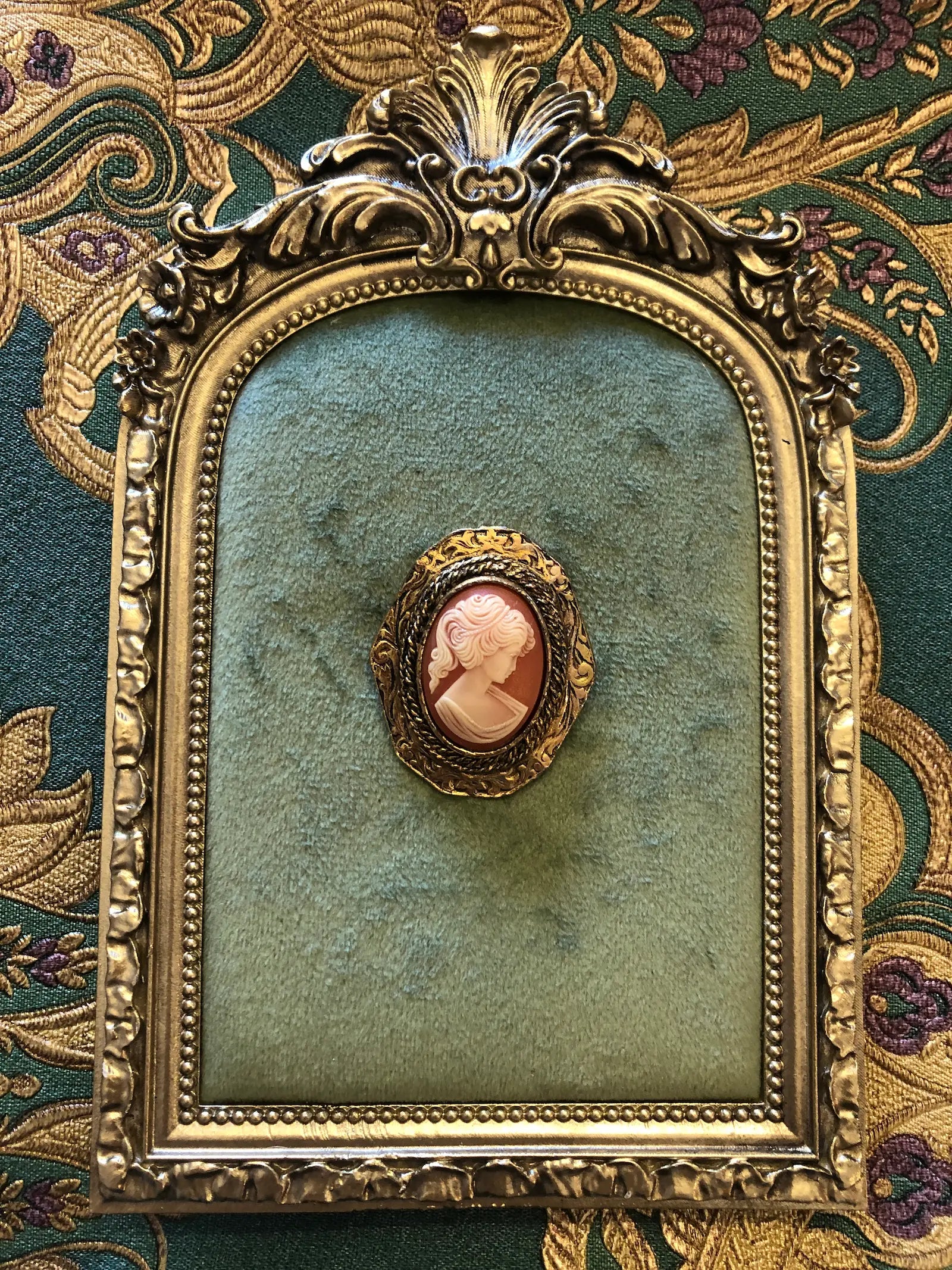 An ornate vintage-style gold frame with a green velvet center, featuring a Broochella Antico Peri oval cameo brooch pinned in the middle, set against a patterned green and gold fabric background.