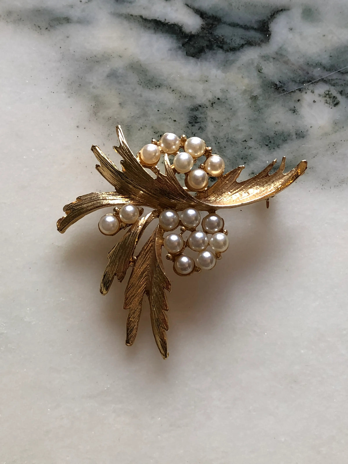 Broochella Antonella Vintage Pearl Leaf Brooch featuring clusters of ivory faux pearls amongst sculptural textured gold-tone leaves on a cream and grey marble background.