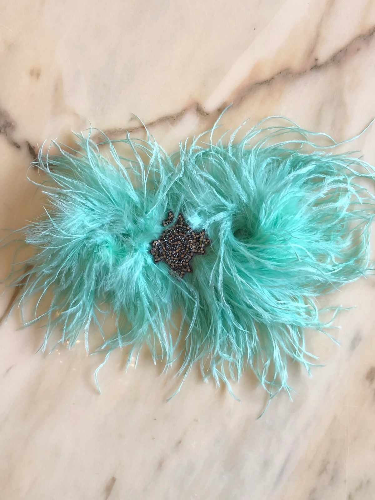 Broochella handmade Aqua Feather Bow Brooch featuring dramatic ostrich feathers and an ornate jewelled centre, an oversized designer statement fashion accessory photographed on a white marble background.