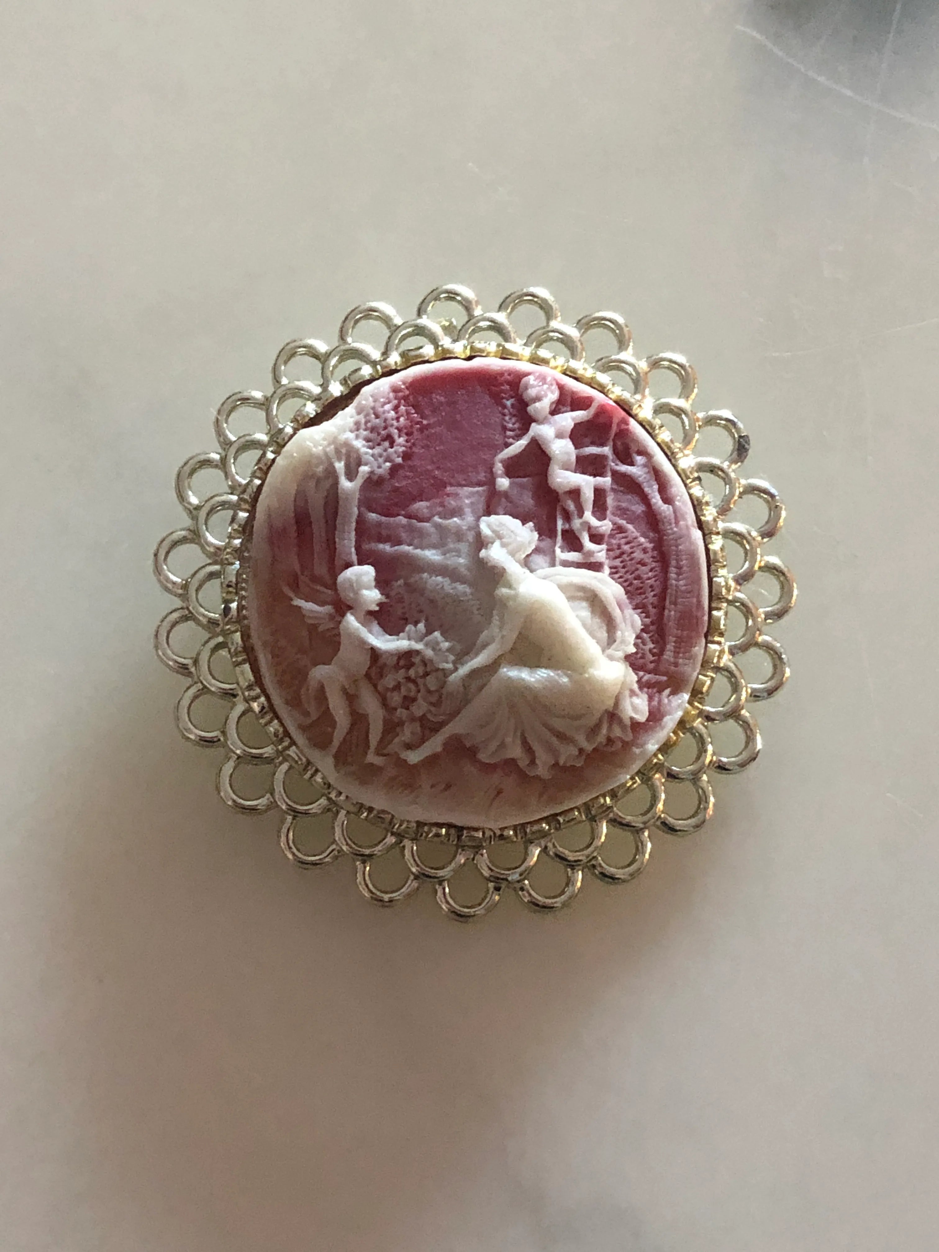 Broochella Arcadia round vintage cameo brooch in pink and ivory, with an ornate, round silver frame. The background is white marble.