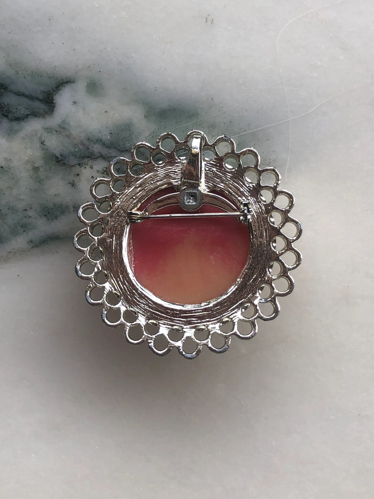 The back view of Broochella Arcadia round vintage cameo brooch in pink and ivory, with an ornate, round silver frame. The background is white marble.