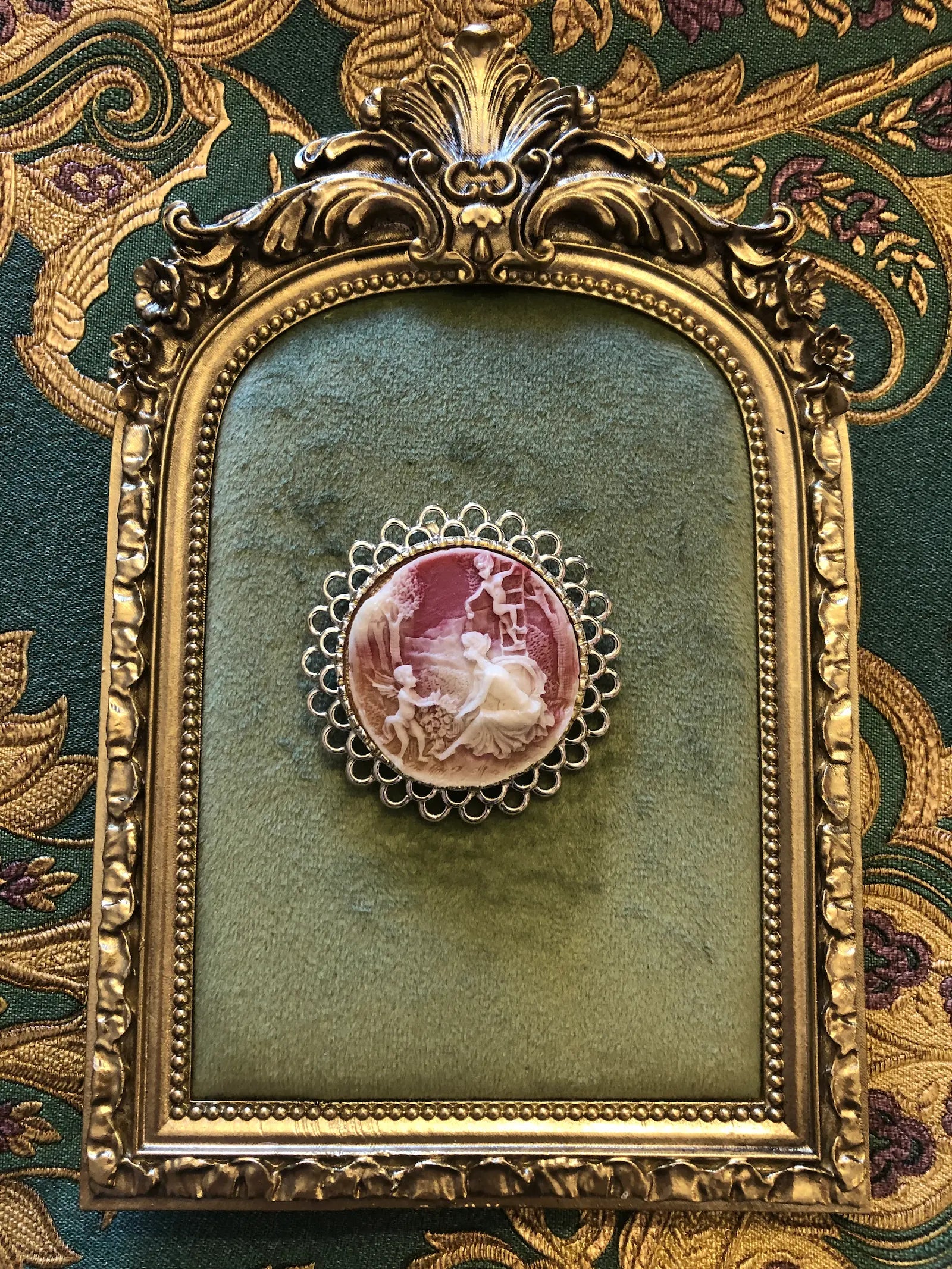 Broochella Arcadia round vintage cameo brooch in pink and ivory, with an ornate silver frame. It is pinned to a green velvet tray with a gold frame. The background is green and gold damask wallpaper.