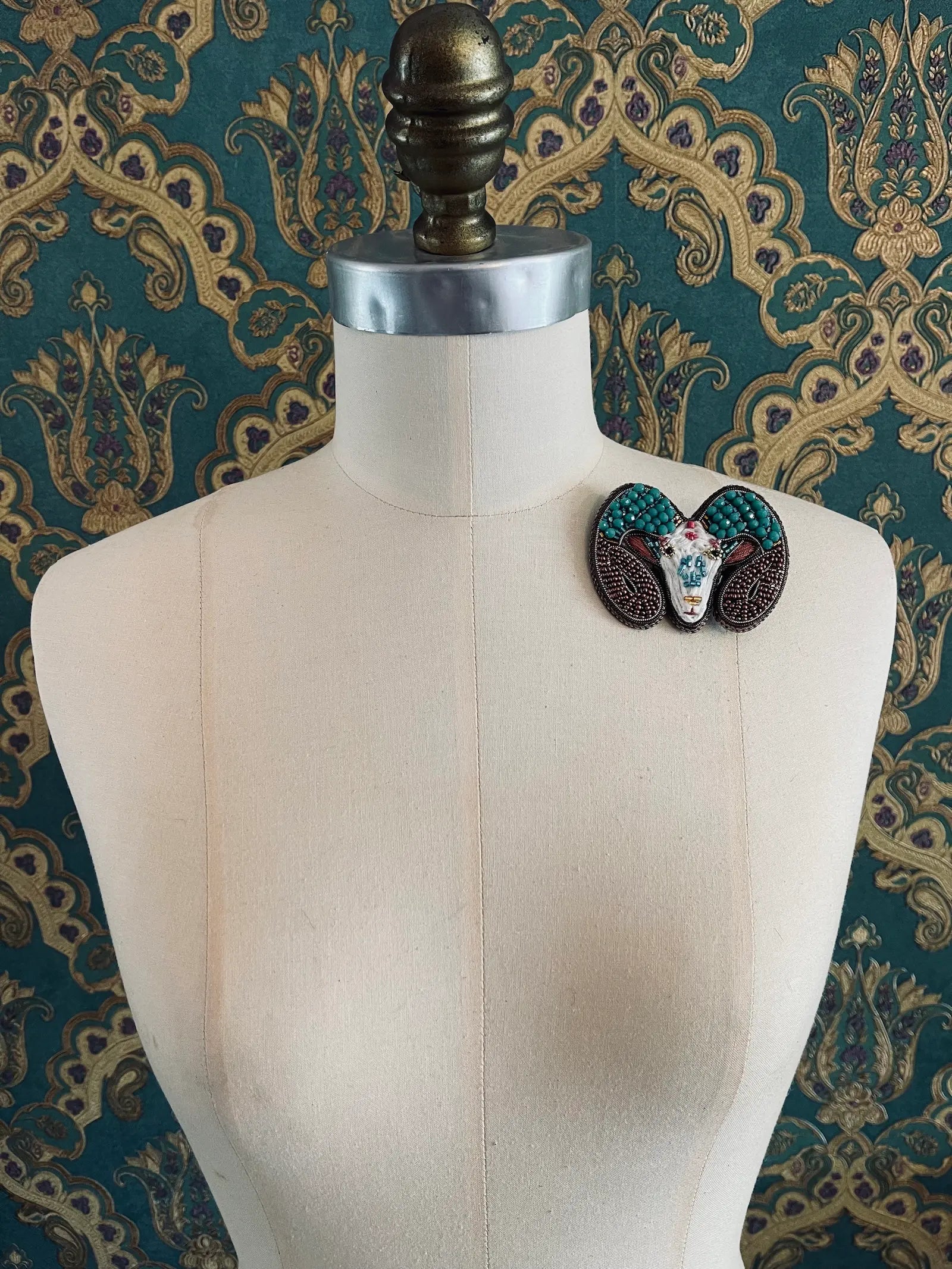 Broochella Aries hand beaded designer zodiac brooch. Styled on a mannequin. The background is green and gold damask wallpaper