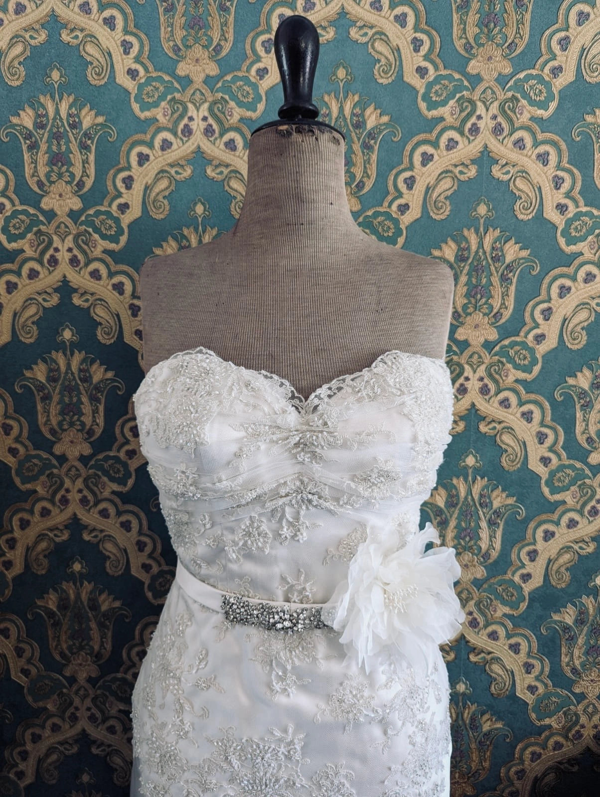 Broochella aubrieta bridal  flower brooch in ivory. Pinned on a mannequin. The mannequin is dressed in a white, beaded, strapless wedding dress. The background is green and gold damask wallpaper.