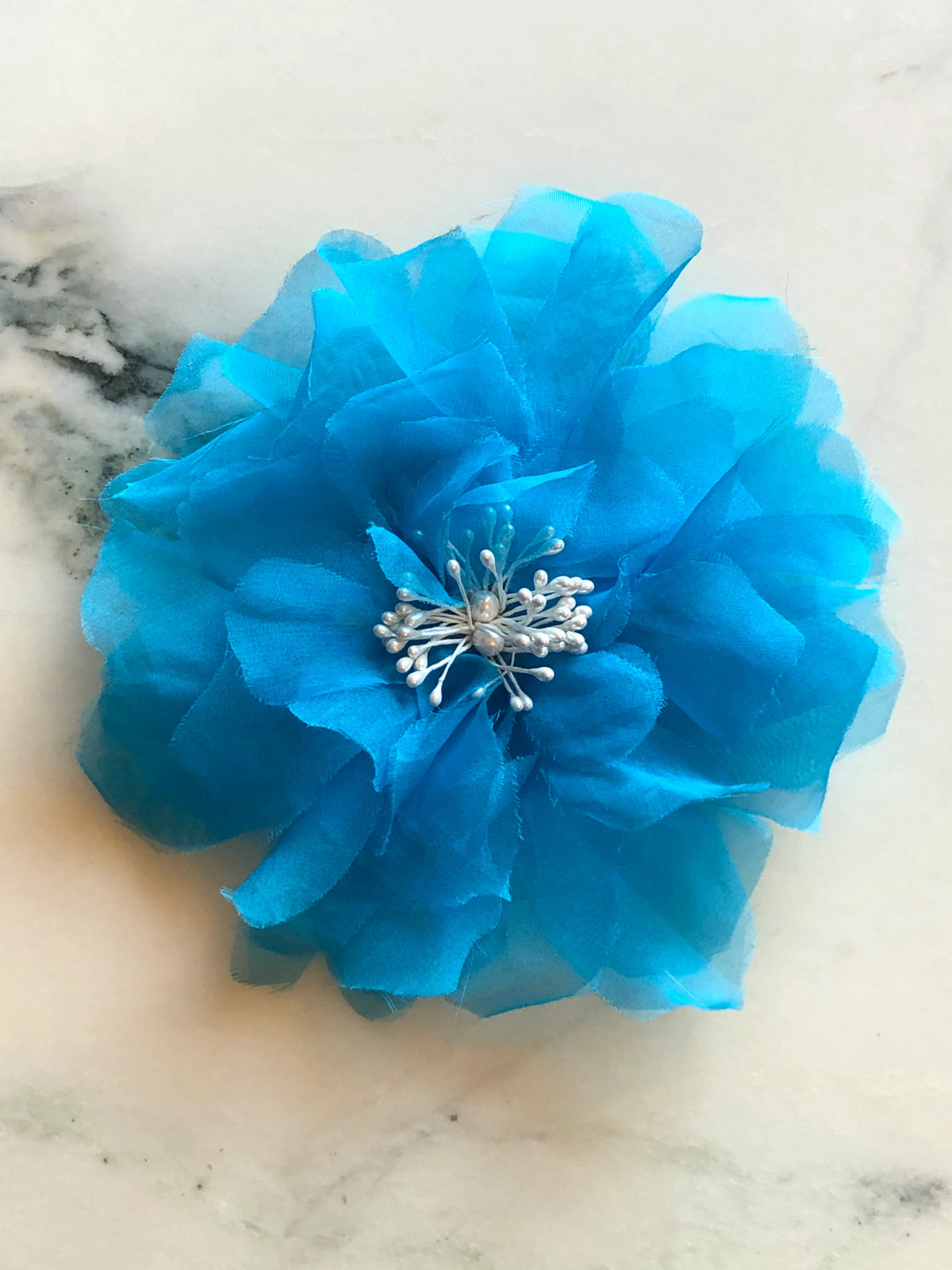 Broochella Aubrieta Flower Brooch Pin in vivid blue, featuring layered sheer petals and a delicate pearl-effect centre. Pictured on a blue marble surface