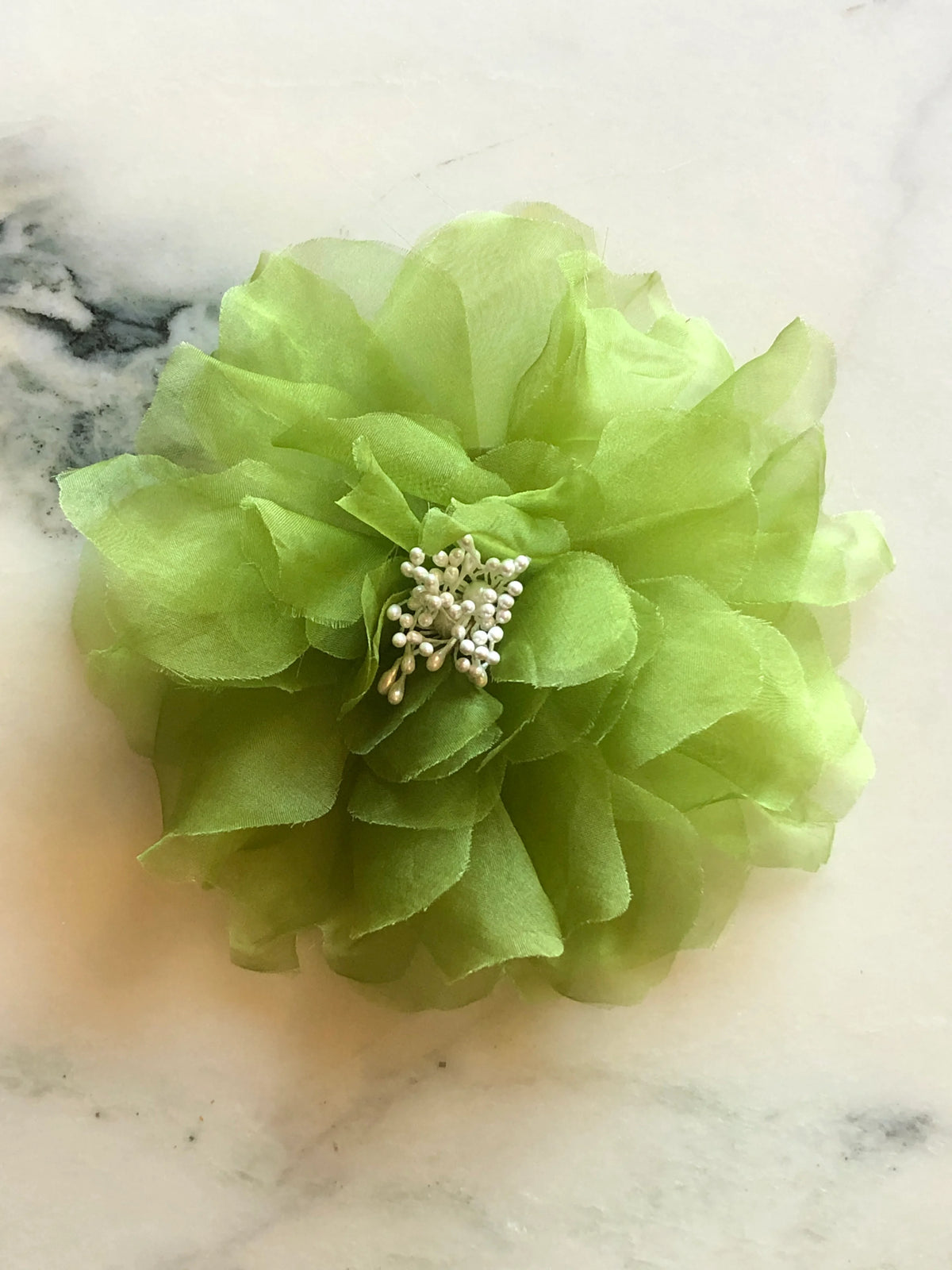Broochella Aubrieta Flower Brooch Pin in chartreuse green, featuring layered sheer petals and a delicate pearl-effect centre. Pictured on a white marble surface