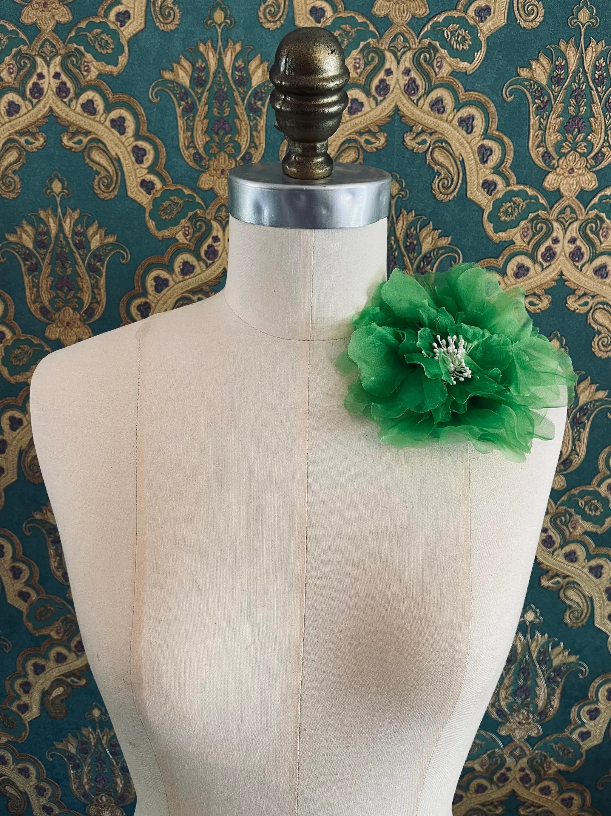 Broochella Aubrieta handmade fabric flower brooch in green. Pictured on a mannequin. The background is green and gold damask wallpaper