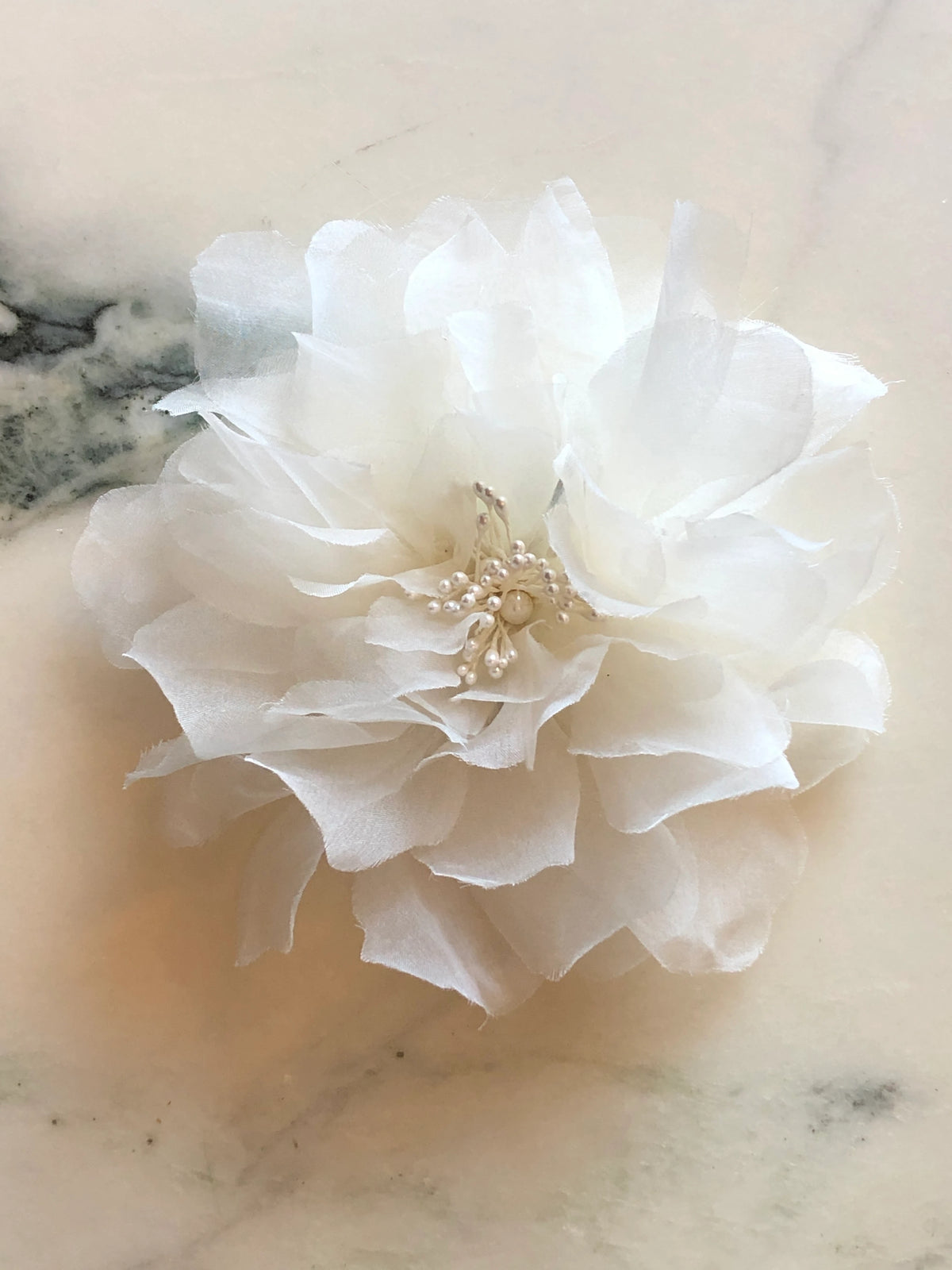 Broochella Aubrieta Flower Brooch Pin in ivory white, featuring layered sheer petals and a delicate pearl-effect centre. Pictured on a white marble surface