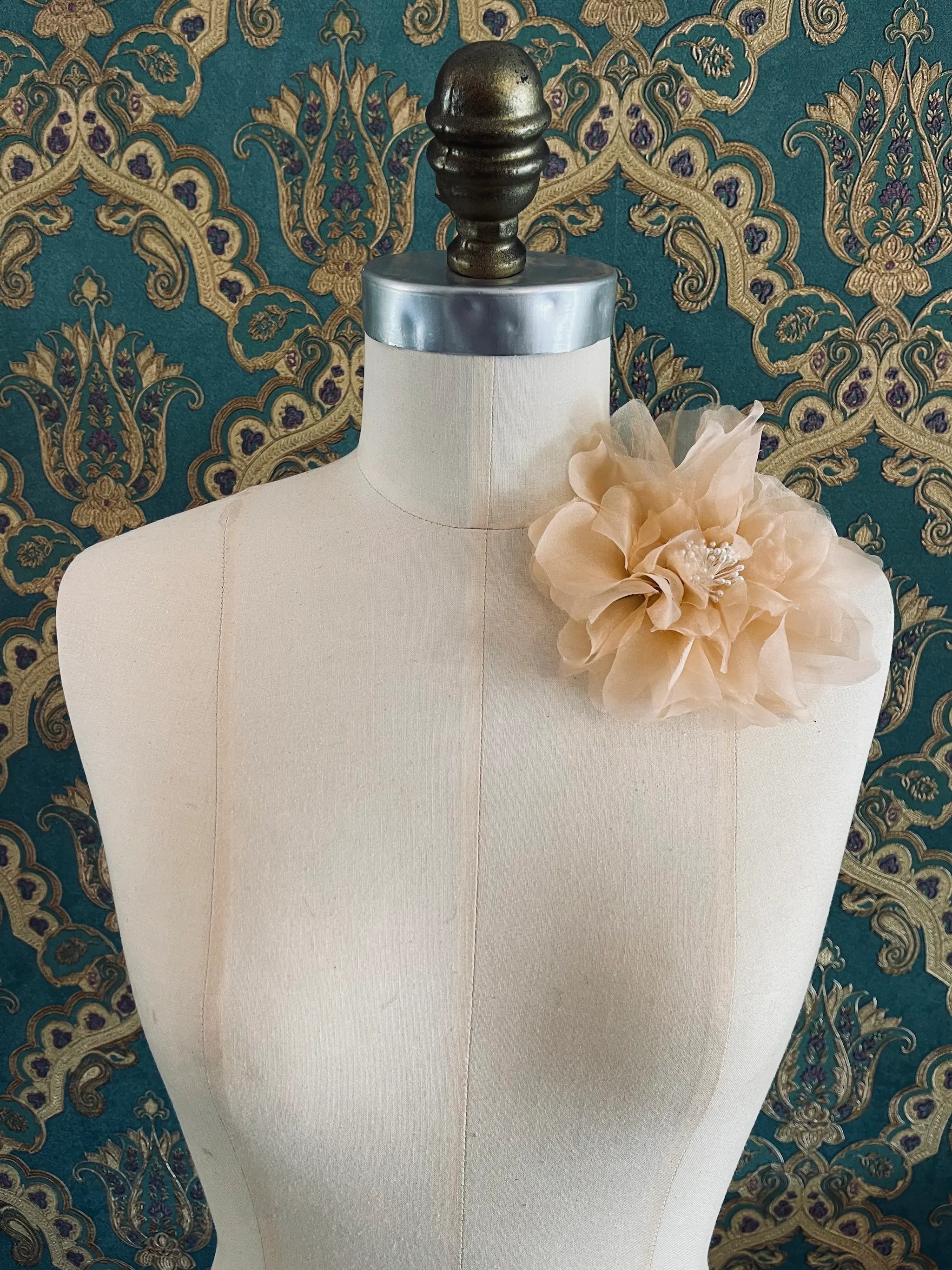 Broochella Aubrieta handmade fabric flower brooch in peach. Pictured on a mannequin. The background is green and gold damask wallpaper