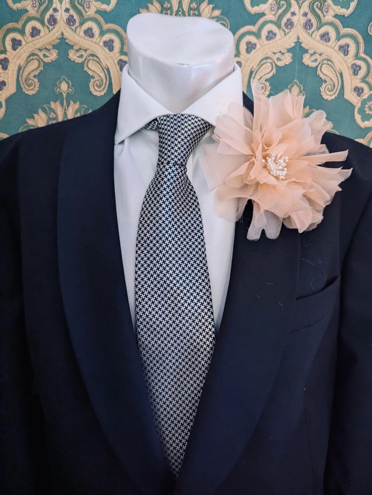 A black suit jacket displayed on a white mannequin torso, styled with a white dress shirt, a patterned grey and white tie, and a Broochella men's apricot Aubrieta flower brooch against a teal and gold damask wallpaper background.