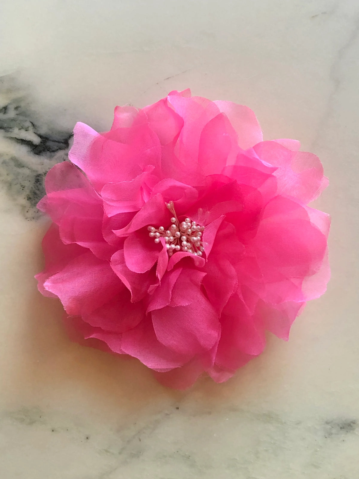 Broochella Aubrieta Flower Brooch Pin in vibrant pink, featuring layered sheer petals and a delicate pearl-effect centre. Pictured on a white marble surface