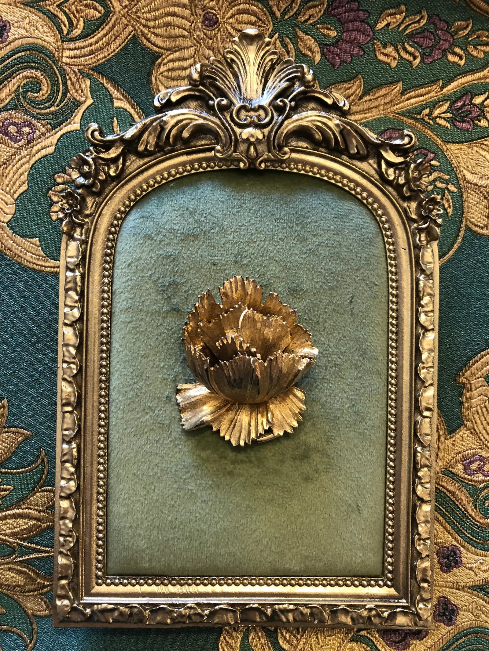 Broochella Aureo vintage gold tone metal flower brooch.  It is pinned to a green velvet tray with an ornate gold frame. The background is green and gold damask wallpaper. 