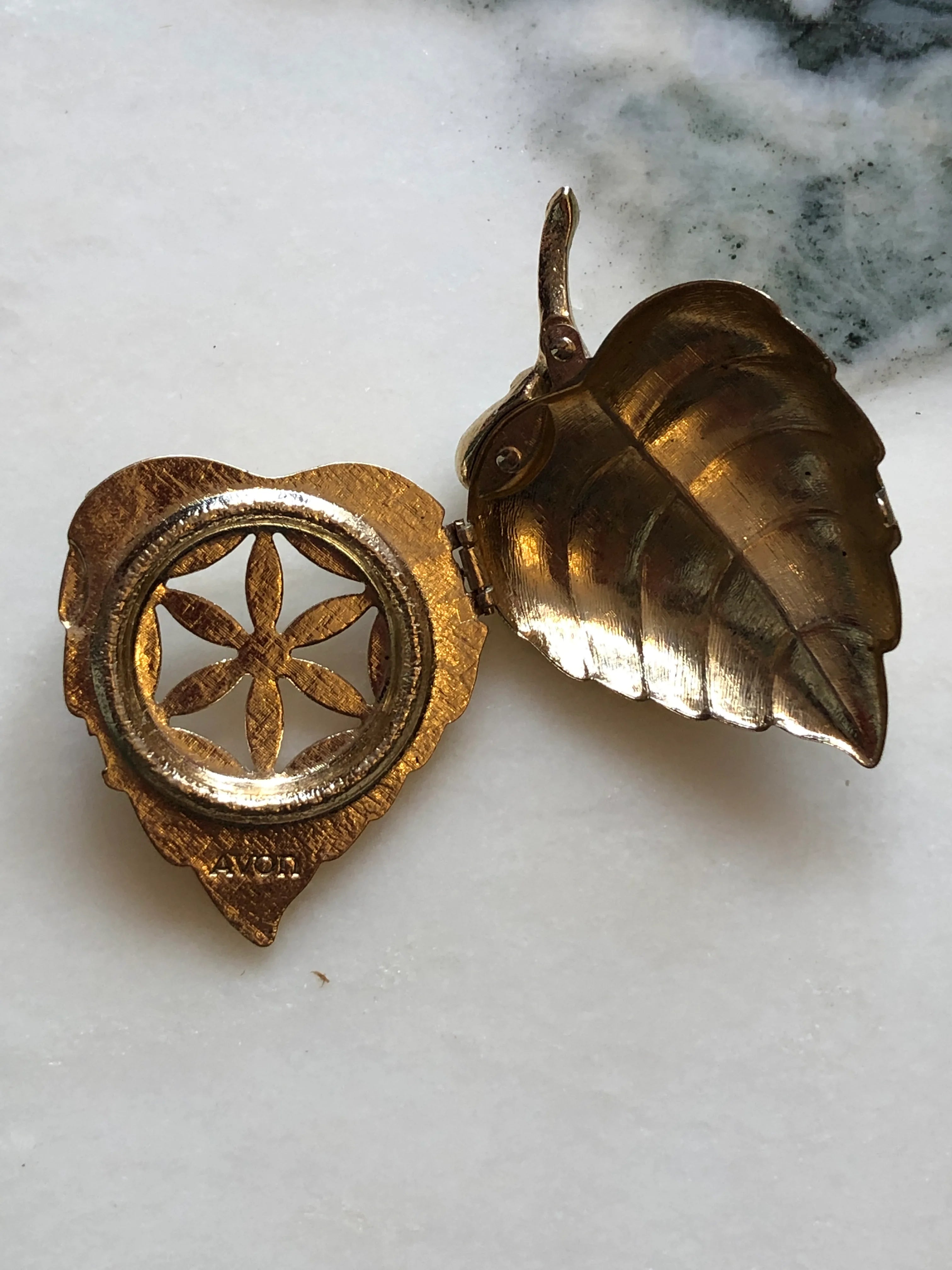 Broochella Avon vintage gold-tone leaf perfume glacé brooch with engraved detailing and a faux pearl accent. It is open, showing the inside of the brooch. The background is white marble