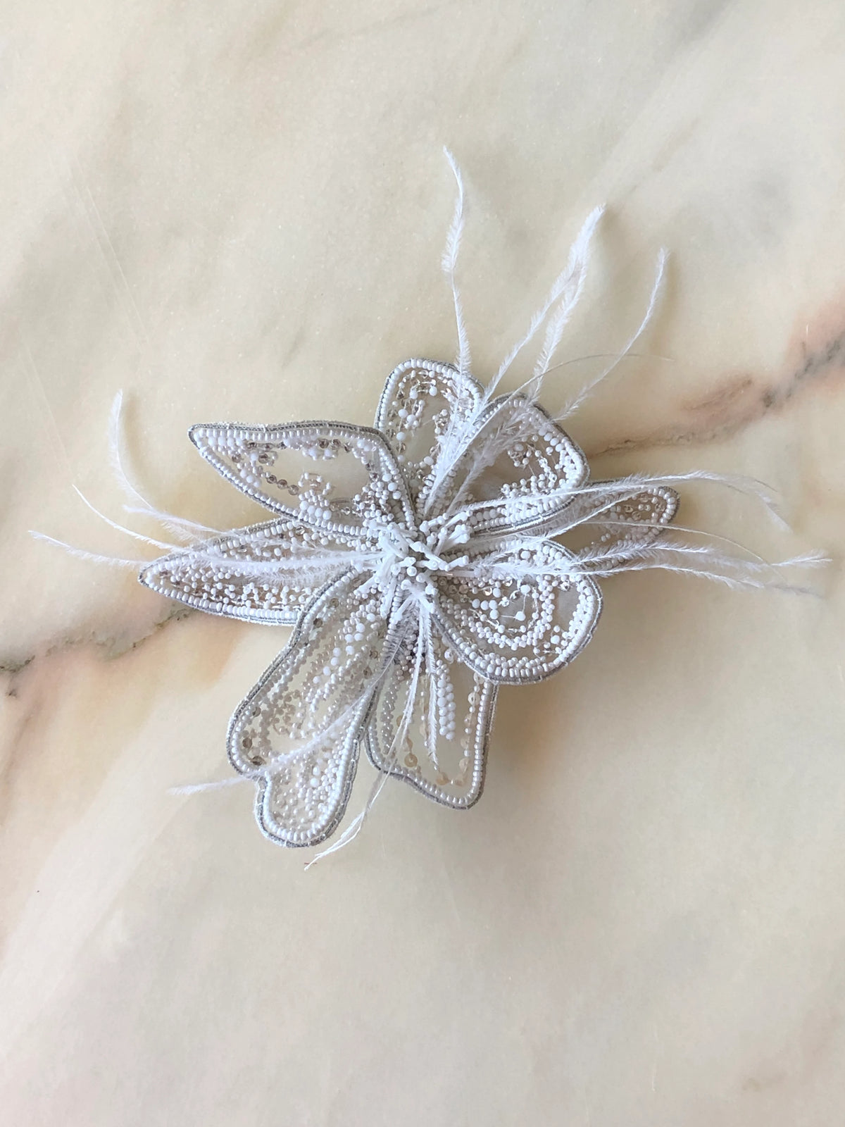 Broochella Men's Azalea Flower Bridal Brooch Small in white, featuring delicate beaded and sequinned petals with soft white feather accents, photographed on a cream marble background.