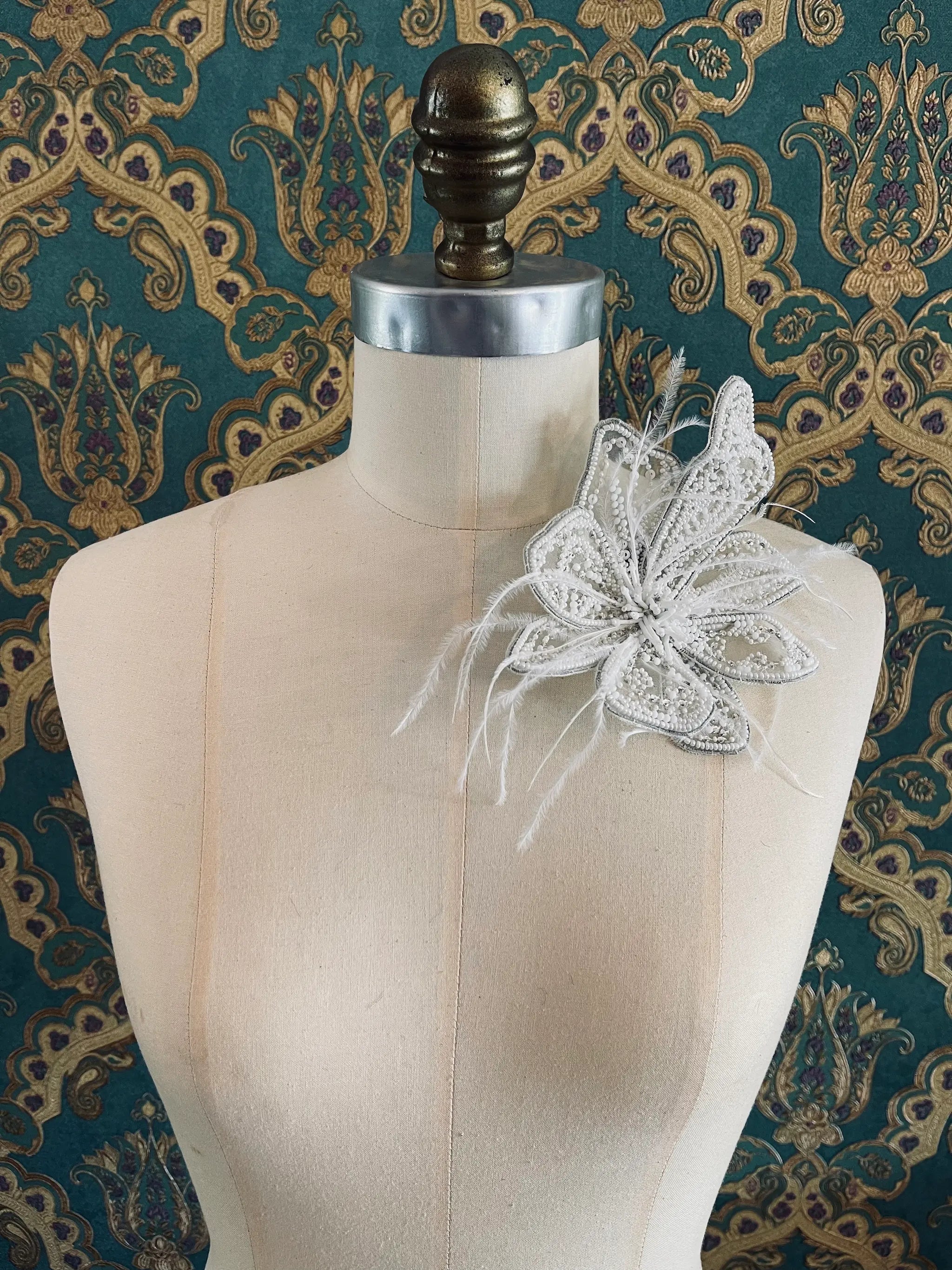 Broochella Azalea hand beaded flower brooch in white. Pictured on a mannequin. The background is green and gold damask wallpaper