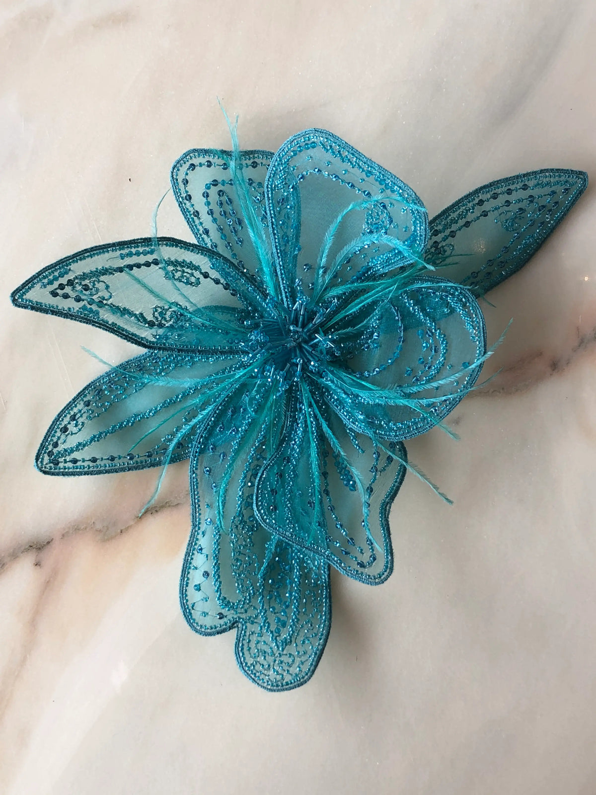 Broochella Azalea Large handmade designer flower brooch in sheer turquoise organza, intricately hand-beaded with tonal turquoise sequins and beads and finished with delicate feather details, photographed on cream marble with natural grey veining.