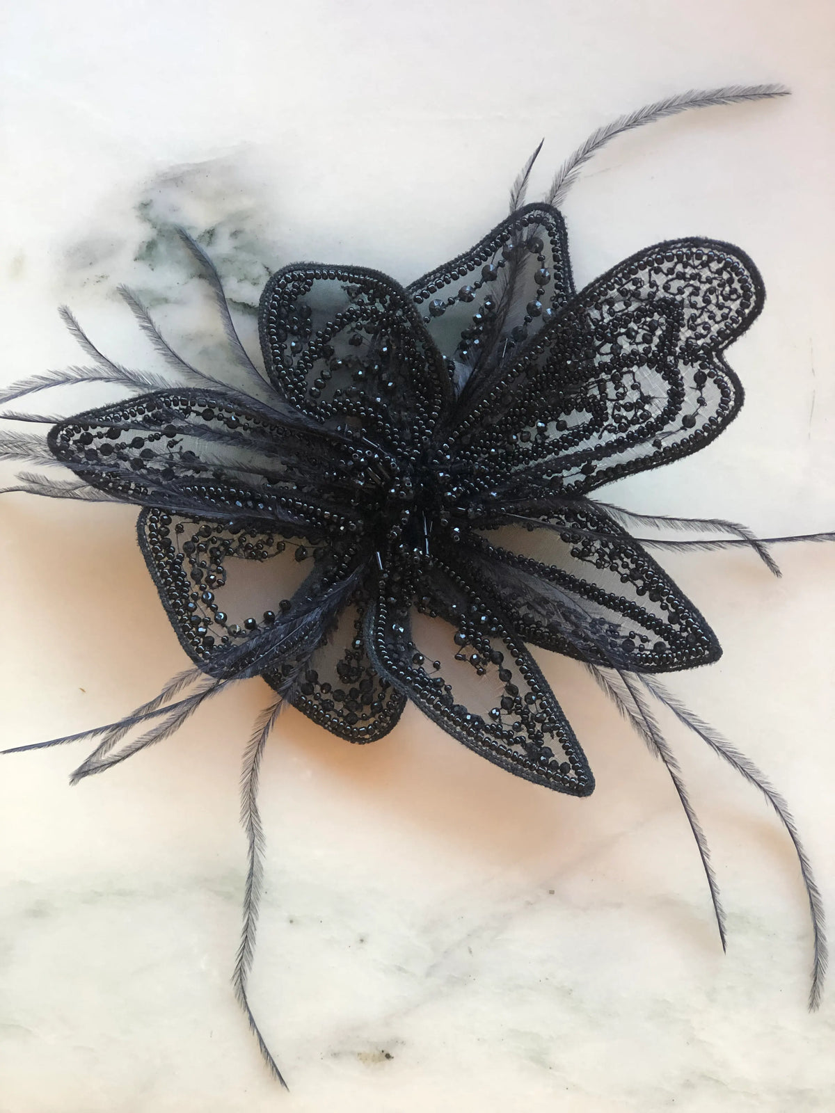 Broochella Azalea Flower Hand-Beaded Brooch Pin - Small in black, featuring intricate black beadwork on sheer sculptural petals with delicate black feather accents on a cream marble background.