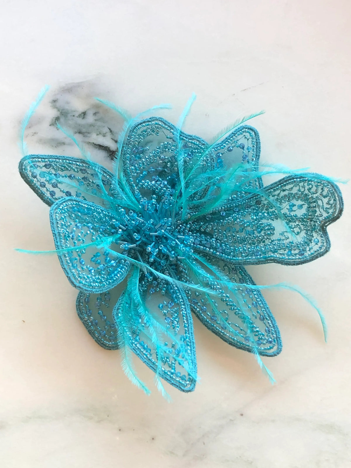 Broochella Azalea Flower Hand-Beaded Brooch Pin - Small in bright turquoise blue, featuring intricate tonal beadwork on sheer sculptural petals with delicate blue feather accents on a cream marble background.