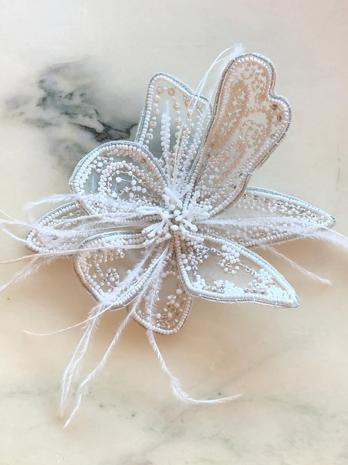 Broochella Azalea Flower Hand-Beaded Brooch Pin - Small in white, featuring intricate white and pearl-toned beadwork on sheer sculptural petals with delicate white feather accents on a cream marble background.