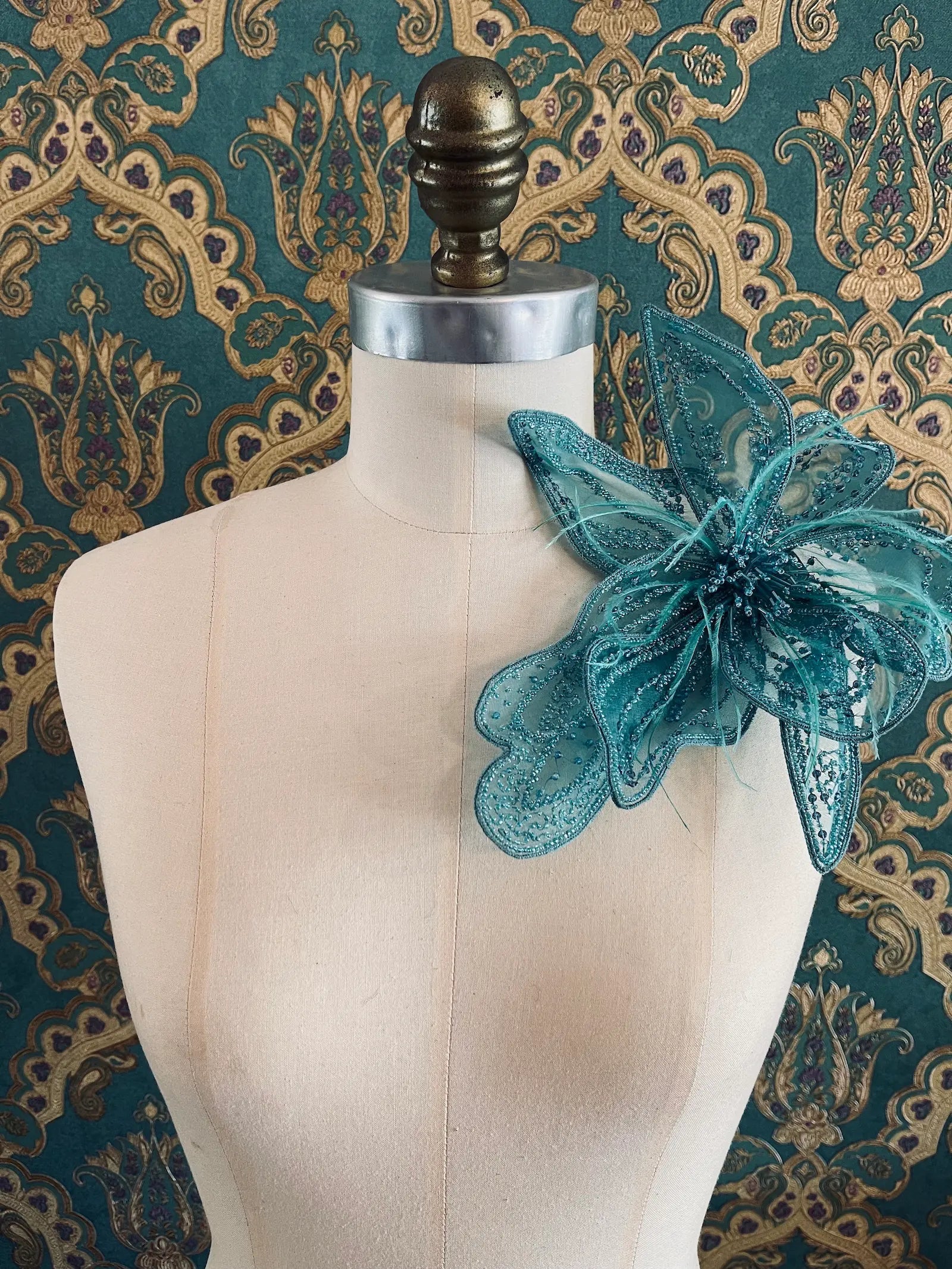 Broochella Azalea large handmade hand beaded flower brooch in blue. The brooch is pinned to a mannequin. The background is green and gold damask wallpaper.
