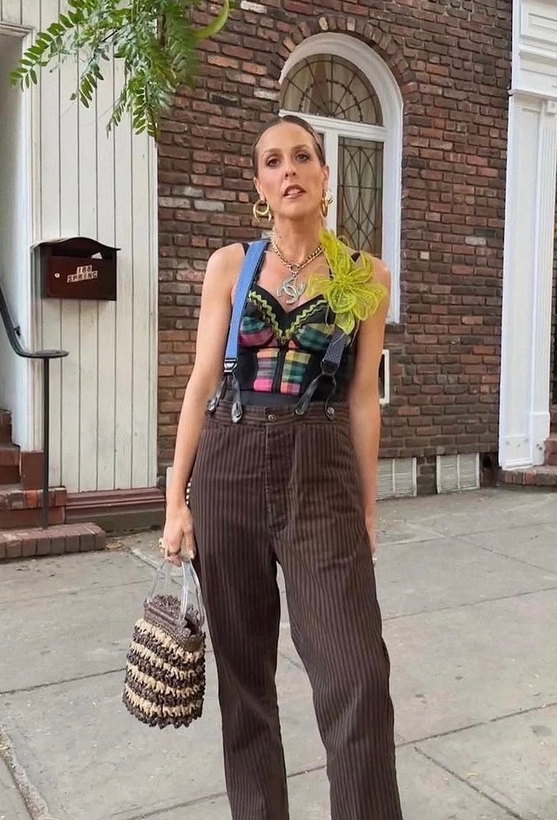 A woman takes a selfie while wearing a black and green patterned top, a blue strap bag, brown pinstriped pants, and gold jewelry, sitting on stone steps in front of a storefront window. She is wearing the Broochella Azalea large, hand beaded, chartreuse flower brooch.