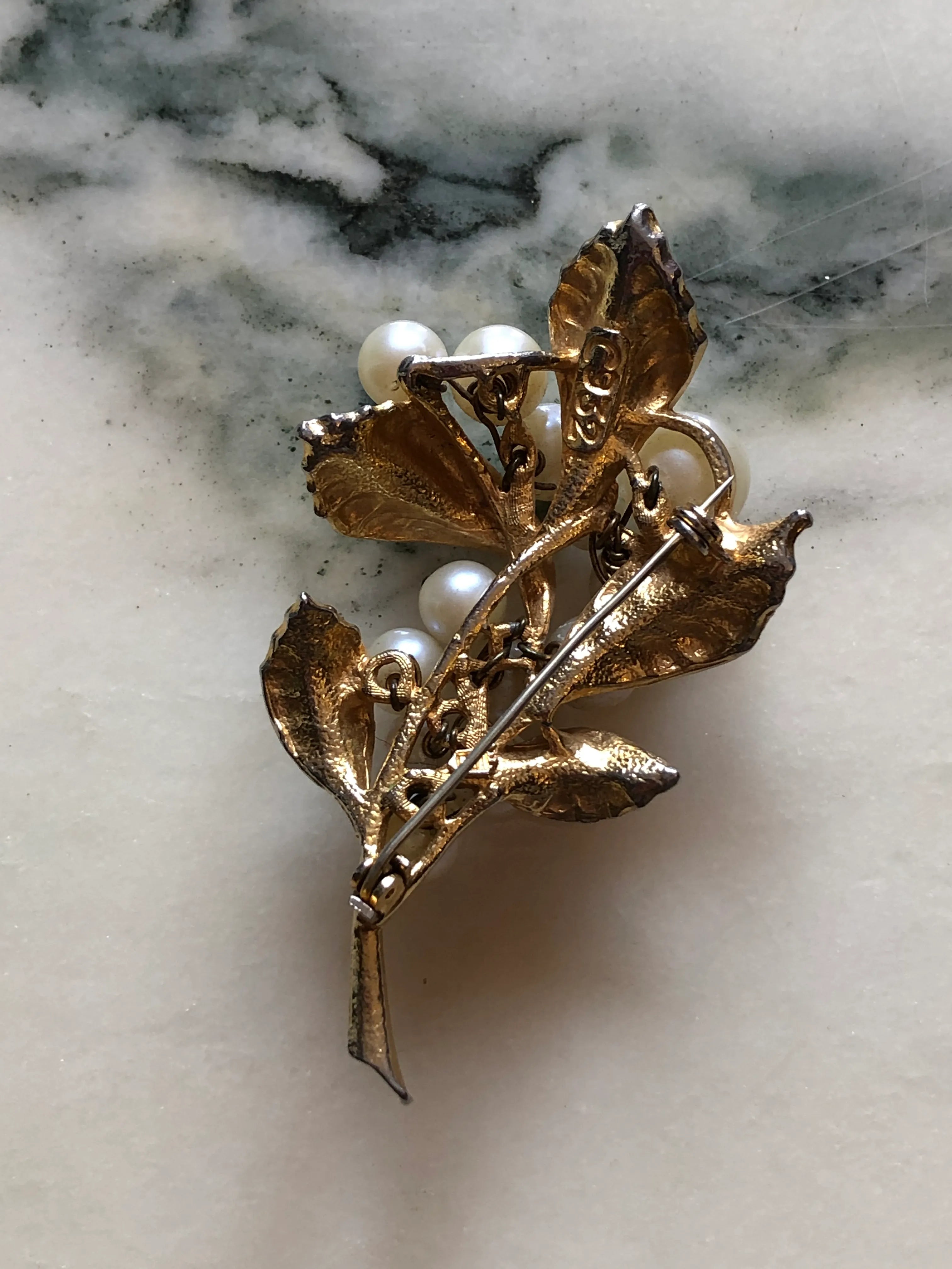 The back view of Broochella Bacche di Perla vintage gold-tone botanical brooch with textured leaves and graduated faux pearl clusters. Pictured on a white marble surface.